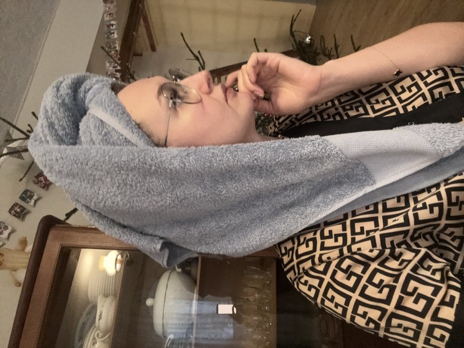 The image captures an indoor scene in Lezhnevo, Russia. A female subject is depicted in profile from the chest up, facing right. She wears eyeglasses with thin, round frames and has her hair wrapped in a light blue terry cloth towel, forming a turban-like shape. Her garment features a repeating black geometric pattern on a light-colored background. Her right hand is positioned with fingers curled against her cheek and chin, and her thumb beneath her jawline. A bracelet with two dark beads is visible on her right wrist.
To the left of the subject, a wooden display cabinet with glass doors is visible. Inside, stacks of white ceramic dishes, including a lidded tureen, are arranged on shelves. A small, illuminated lamp with an opaque shade is present within the upper section of the cabinet, and clear glassware is visible on a lower shelf. Above the cabinet, small rectangular decorations are affixed to the wall, along with a light-colored figurine. To the right of the subject, a section of a Christmas tree or large branches, adorned with at least one silver bauble, is visible against a light-colored wall.