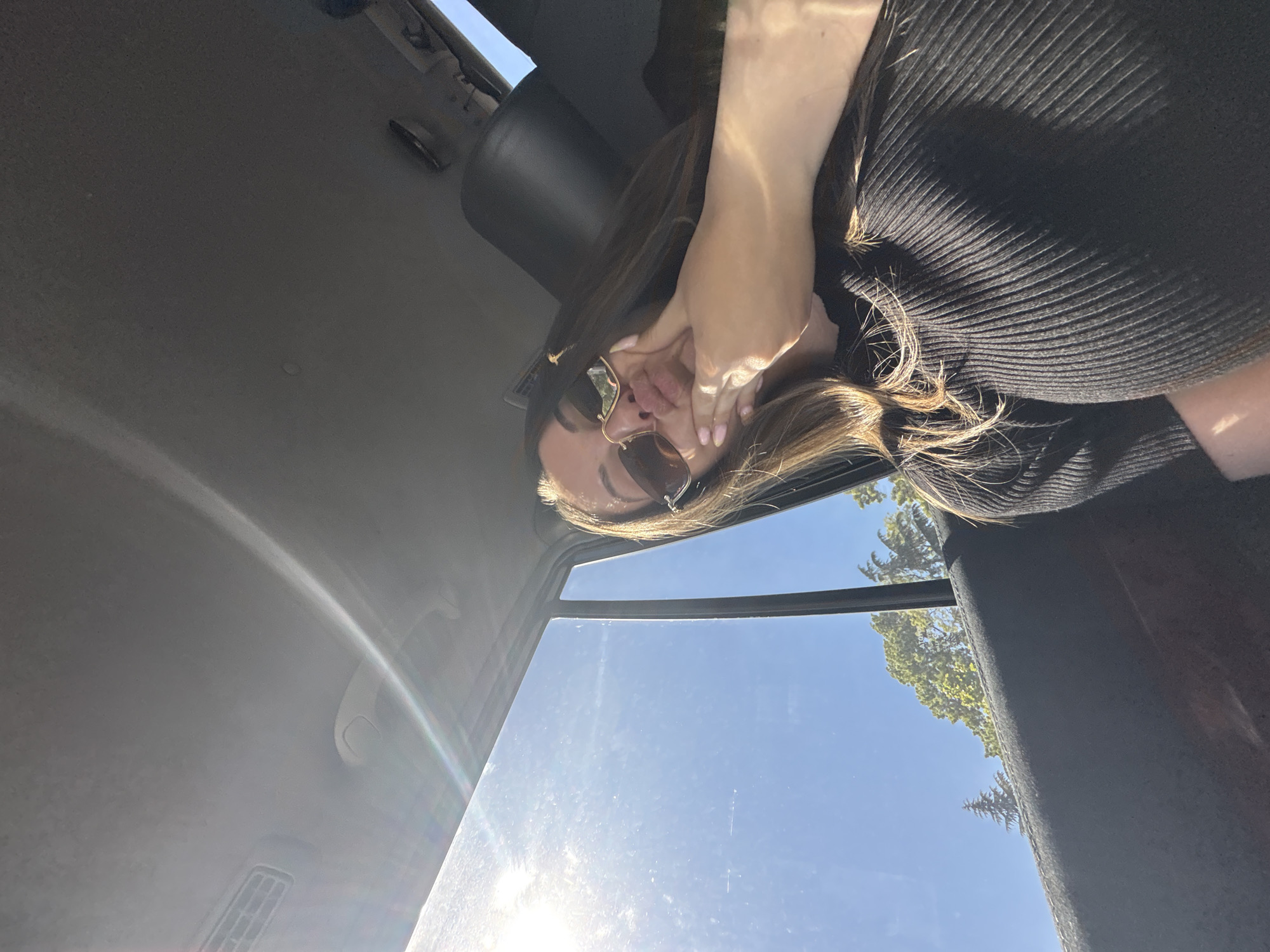 A low-angle shot from inside a vehicle looking towards the sky through the car window, shows a woman wearing sunglasses and a black, ribbed top. She is holding her hand up to her face. Outside the window, there are trees and a clear, blue sky. The location cannot be pinpointed to the city of Burgas, Bulgaria without additional information or landmarks visible in the image. The interior of the car includes the ceiling, driver's seat, and part of the dashboard visible due to the camera angle.