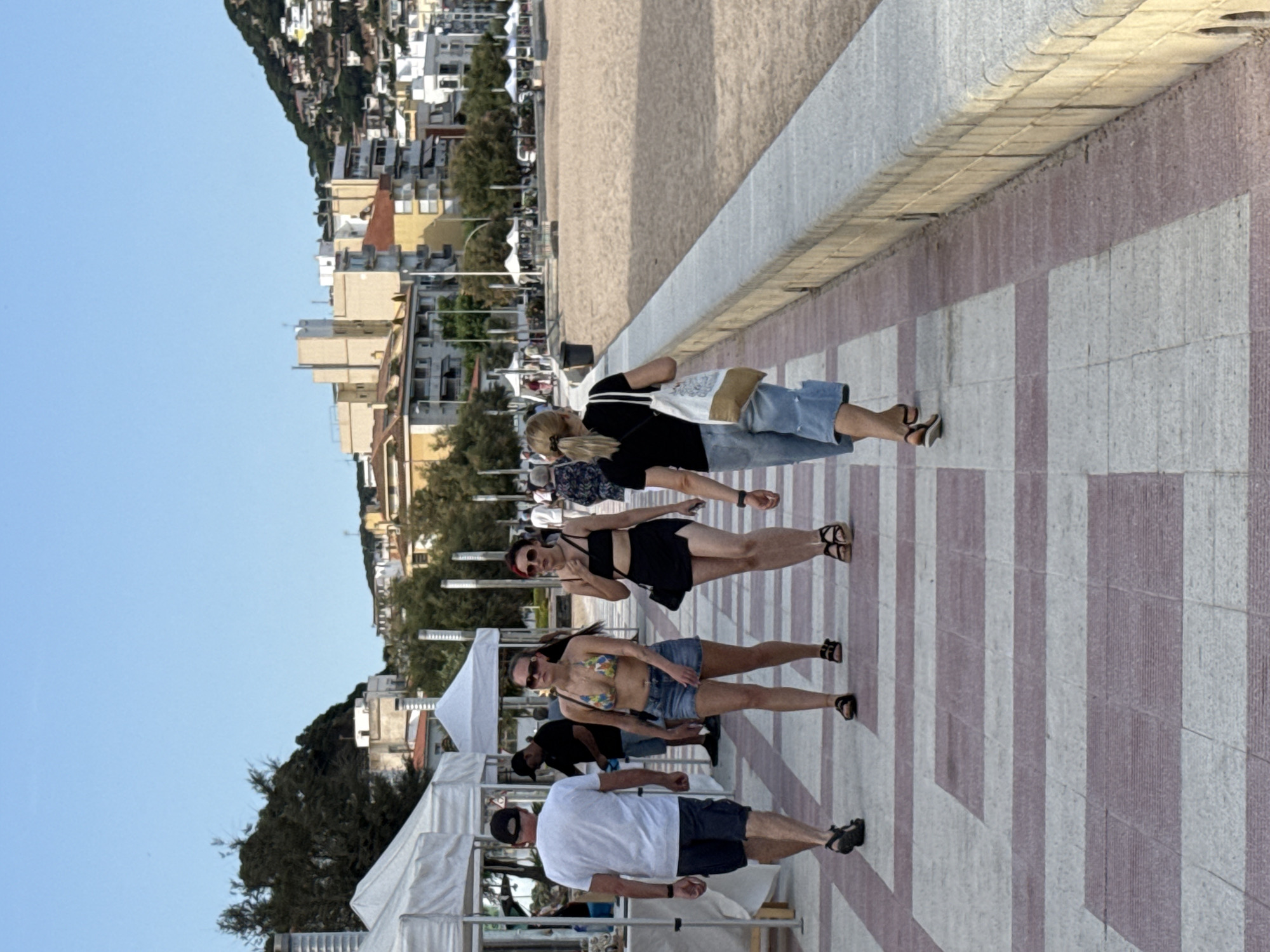 This image depicts a sunny day scene in Blanes, Spain, likely in the late afternoon or early evening given the warm lighting. The foreground is dominated by a patterned tiled walkway, with people strolling along it. The subjects are a mix of individuals and small groups, all casually dressed, suggesting a relaxed atmosphere.
Several individuals are clearly visible. One woman is wearing a black bikini top and a dark skirt with sandals. Another woman is in a colorful bikini top and denim shorts, also wearing sandals. Further down the walkway, a man in a white t-shirt and dark shorts, wearing a baseball cap, walks with his back to the camera. Behind him, another man in a black t-shirt and a black baseball cap is visible. A woman with blonde hair, wearing a black t-shirt and denim shorts, walks away from the camera, carrying a white tote bag.
The background reveals a beach with sand, beyond which lies a town with buildings cascading down a hillside. Lush green trees are interspersed with the urban landscape. The clear blue sky suggests fair weather. In the far left of the image, white canopies of market stalls or outdoor seating areas are visible, hinting at a public area or event. The overall mood is one of leisure and a typical day at a coastal town. No visible text is present in the image.