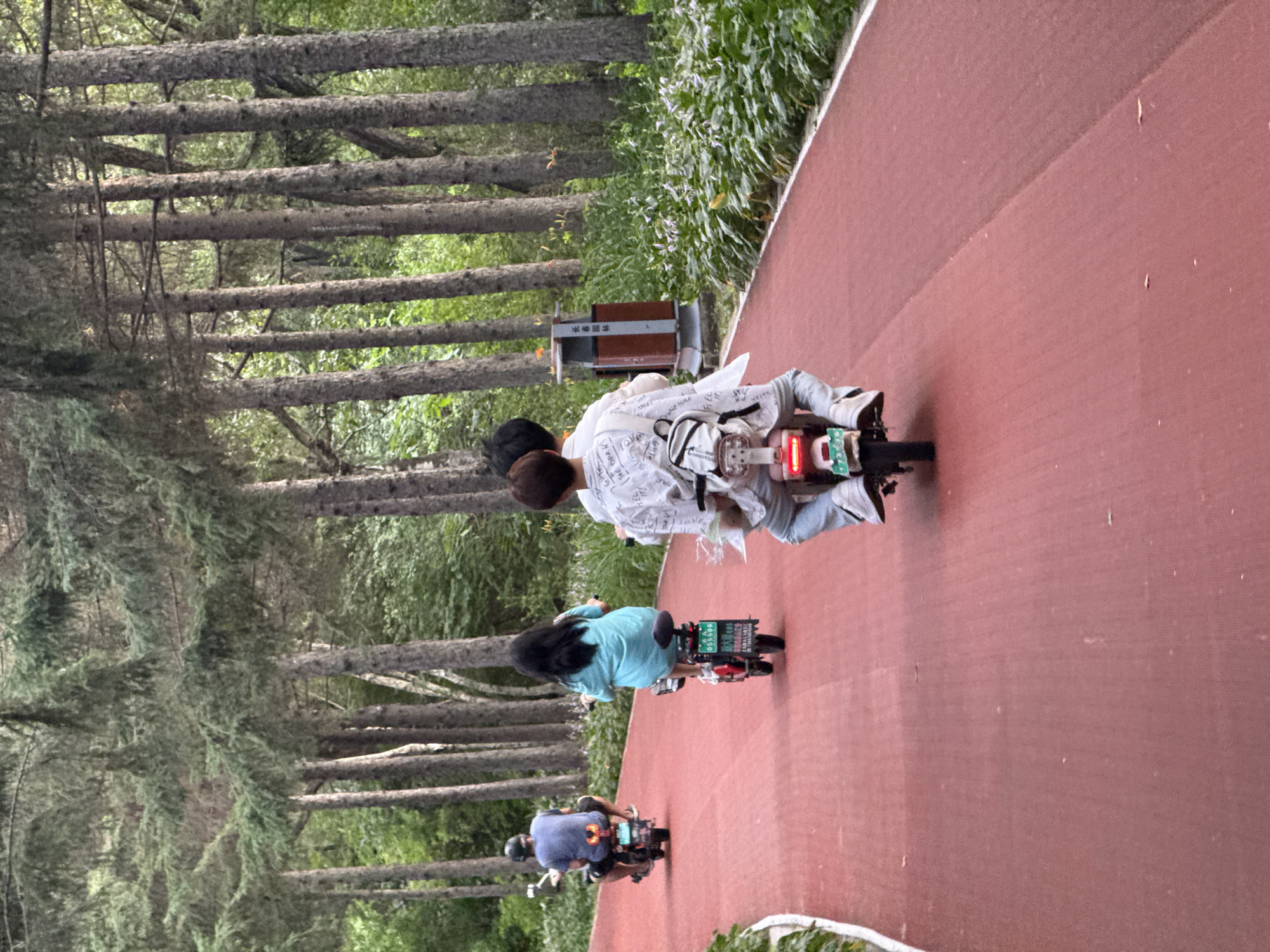 In Changchun City, China, on a red paved pathway lined with tall pine trees, three individuals are riding electric scooters. The leftmost scooter is ridden by a person wearing a blue t-shirt and a helmet. Next, a person in a teal top rides a scooter with a license plate that reads "055506". The third scooter, closest to the viewer, carries two individuals. One is wearing a white patterned shirt and jeans, with a backpack on their back. The other person, partially visible, is also wearing a white shirt. A red and silver trash can is visible behind the scooters, near the edge of the pathway, among lush green foliage. The scene appears to be a park or recreational area.