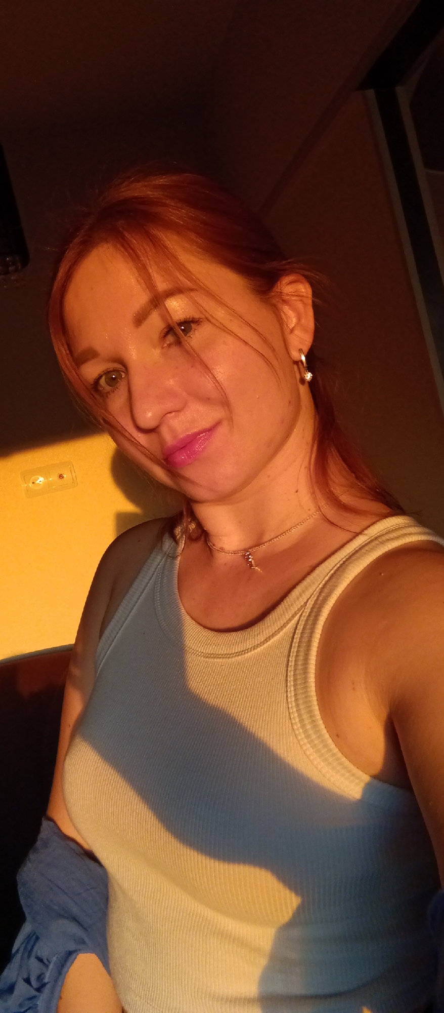 A woman with reddish-blonde hair pulled back is positioned in the foreground of the image, wearing a white tank top and a delicate gold necklace. A patch of bright yellow light illuminates part of her face and the wall behind her, suggesting the presence of a sunset or strong artificial light source. A partially visible electrical outlet is on the wall. The light casts shadows on the woman's clothing and skin. Part of a blue garment is visible on the lower left. The location in the city is unknown.