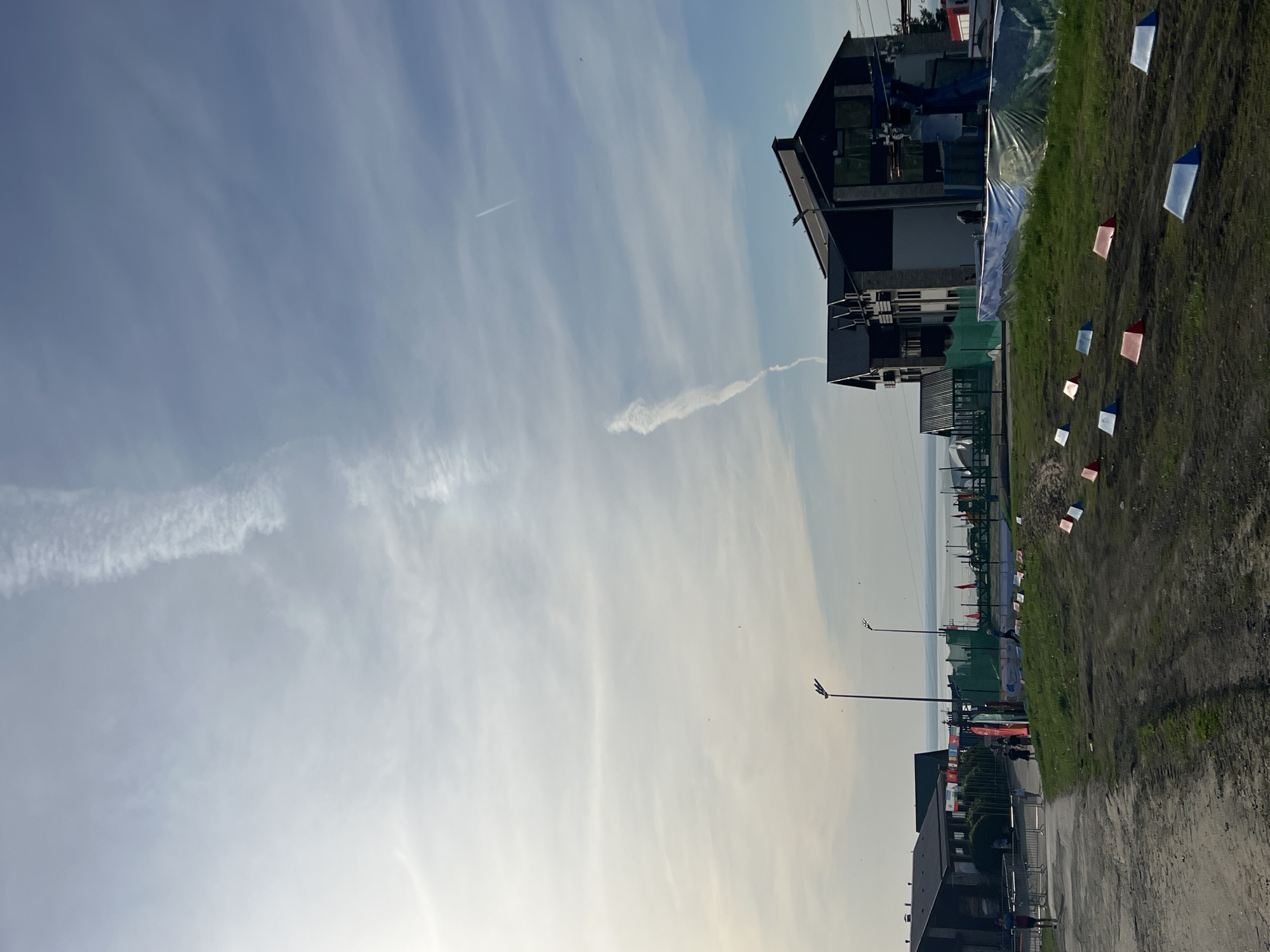 The image presents a partially obscured landscape in Khvalynsk, Russia. A building is visible on the right with partial plastic sheeting visible. A row of red and white flags adorn a patch of grass near the building. Down a row of streetlights, with other various buildings in the background. A contrail is visible against a blue sky background.