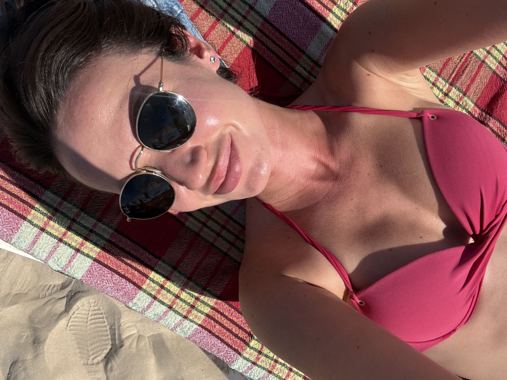 A first-person perspective shot captures a person lying on a beach in Tel Aviv, Israel. The subject, wearing a pink bikini and sunglasses, basks in the sun on a red, yellow, and green plaid blanket. The warm sunlight creates a glowing effect on their skin and casts shadows across the scene. A faint footprint in the sand beside the blanket suggests a recent presence or movement. The image evokes a sense of relaxation and enjoyment of a sunny day at the beach, with the bright sunlight indicating daytime. There is no visible text in the image.