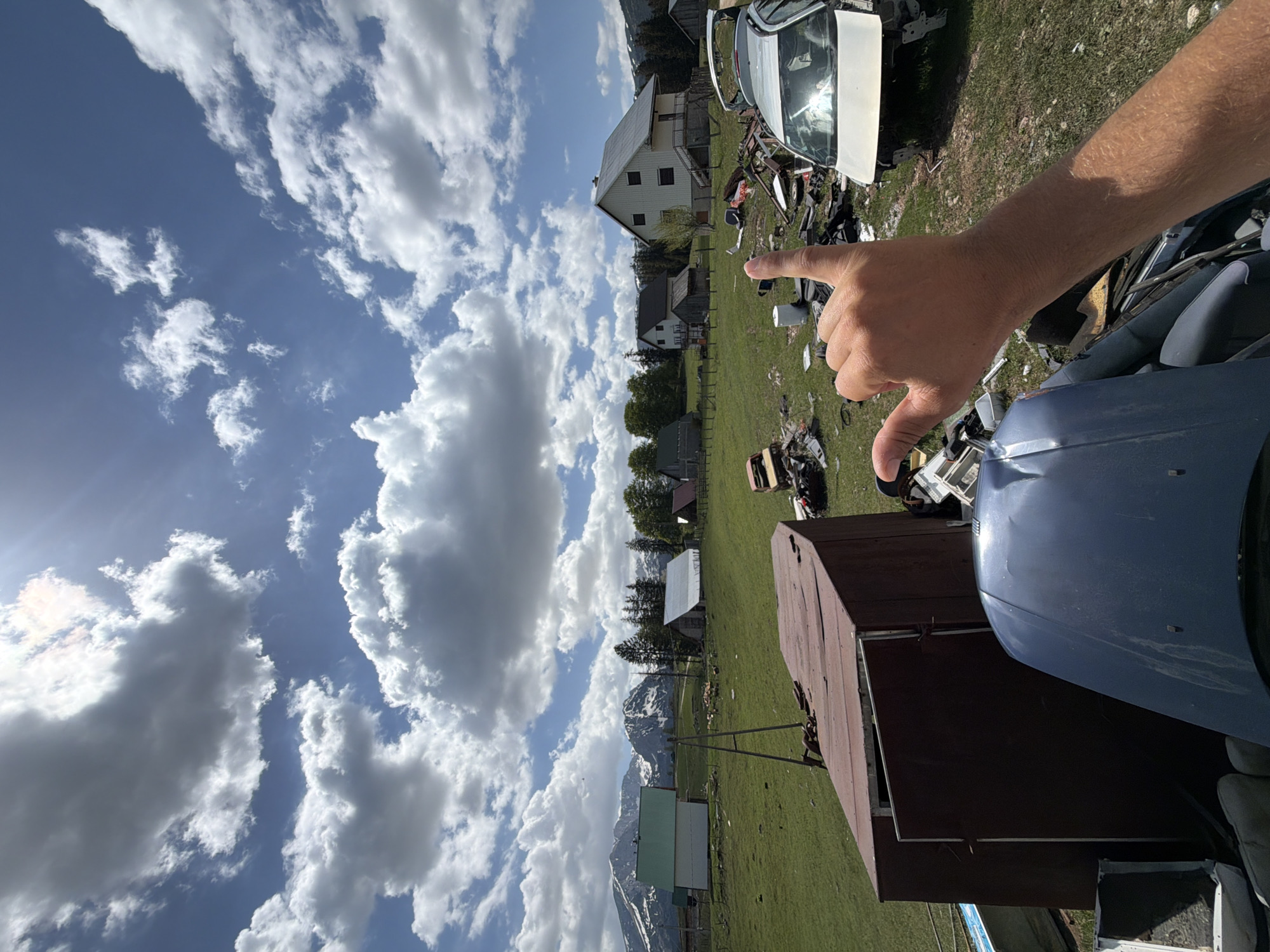 The image shows a rural scene in Žabljak, Montenegro. The sky is bright blue with scattered cumulus clouds. In the foreground, a hand points towards a white building amidst a field littered with scrap metal, presumably a junkyard. The mountains are visible in the background. A partially visible car and shed also appear in the immediate foreground.
