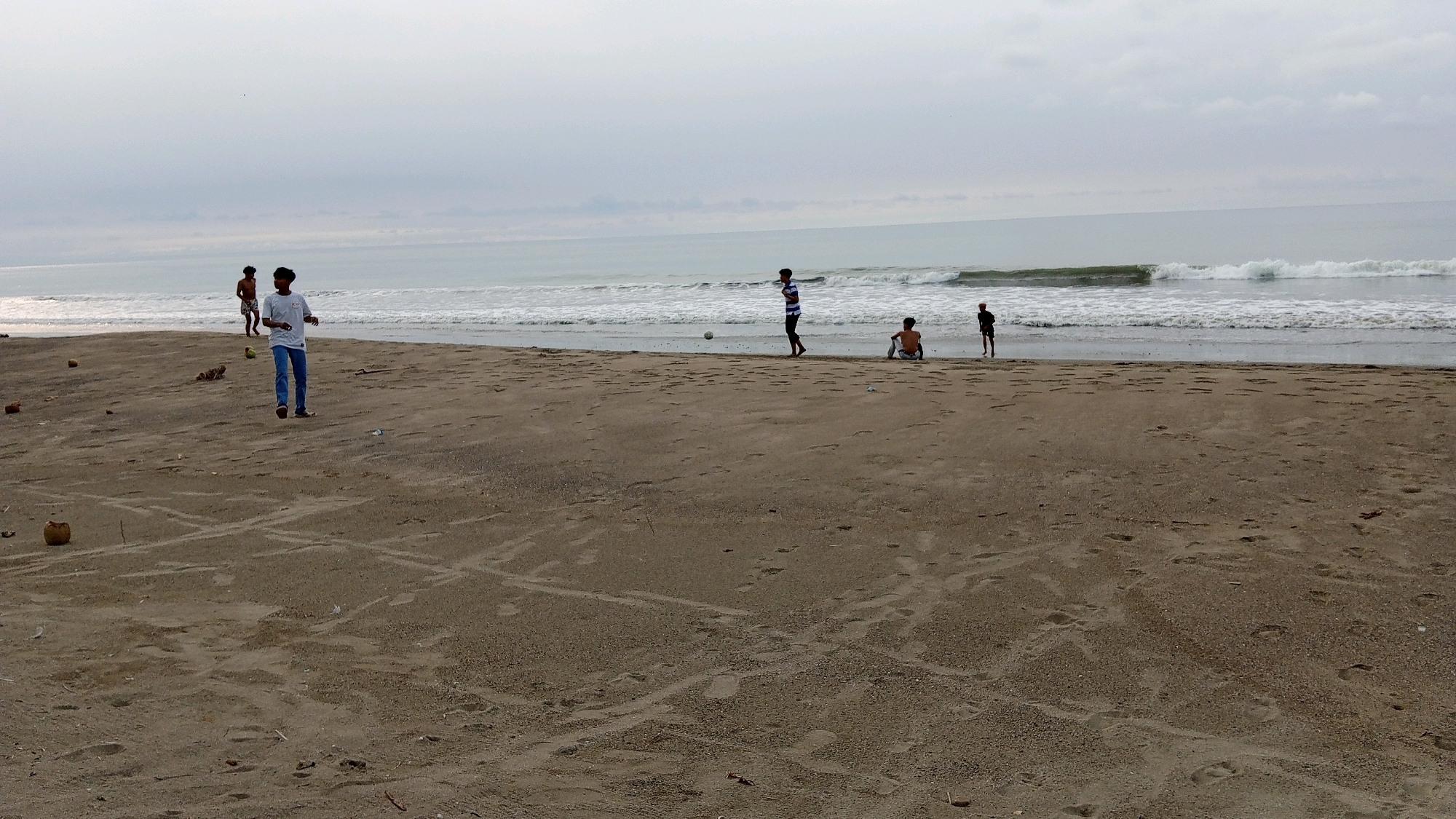 A wide, low-angle, outdoor, daytime, medium shot captures a sandy beach in Kruengjuli Timu, Indonesia. A muted, overcast sky with light gray clouds stretches across the horizon, meeting a calm, gray ocean with gentle waves. The beach itself is predominantly sand, with scattered debris like coconuts and small pieces of trash. Several individuals are present. In the foreground, a lone person walks towards the camera wearing a white t-shirt and blue jeans. Further down the beach, near the water's edge, a group of four young males are scattered. One is in the process of kicking a soccer ball, while two others are sitting or standing near the shoreline. A fourth person is standing slightly further back. The overall atmosphere appears relaxed and leisurely.