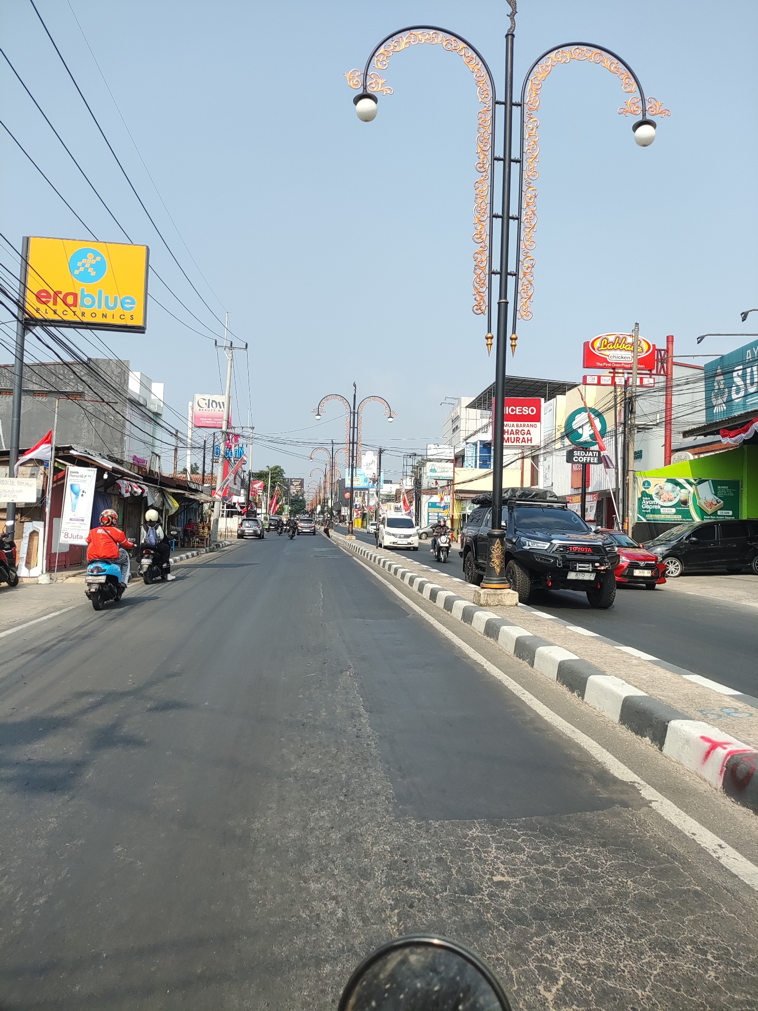 The image depicts a street scene in Majalengka, Indonesia, under a clear blue sky. The viewpoint appears to be from a motorcycle, as indicated by the lower portion of a rearview mirror at the bottom of the frame. The street is paved and lined with businesses on both sides. On the left, a yellow sign for "erablue ELECTRONICS" is visible. Several shops with corrugated metal roofs line the street, with some featuring awnings and signs. Indonesian flags are displayed on some buildings.
The road is moderately busy with vehicular traffic, including motorcycles, cars, and what appears to be a utility vehicle with a roof-top tent. A person in a red jacket and helmet is riding a blue scooter on the left side of the road, followed by another motorcyclist in a white helmet. On the right side of the road, a black Toyota SUV with off-road modifications is prominently parked or moving slowly. Further down the street, other vehicles and pedestrians are visible.
Streetlights with ornate, decorative metalwork are positioned along the right side of the road. Various commercial signs are displayed, including one for "Labbies Chicken," "GLOW BEAUTY NA," and another with text in Indonesian, possibly advertising a sale with "HARGA MURAH" (cheap prices). A green sign shows an image of food, likely a meal, with prices. The road has visible lane markings and a curb with black and white stripes. The pavement shows some signs of wear and tear, with patches of newer asphalt and cracked surfaces. The overall impression is that of a bustling urban street in Indonesia.