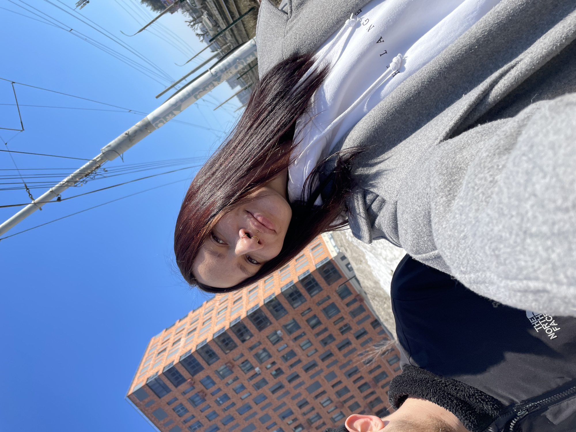 An outdoor scene in Izhevsk, Russia, on a clear, sunny day, likely late winter or early spring due to the presence of dirty snow. The image is a close-up taken from a low angle, focusing on two individuals in the foreground and urban infrastructure in the background.
A female subject occupies the right and center of the frame, facing the camera with a slight smile. She has long, dark reddish-brown hair and a pierced nose, exhibiting both a golden hoop and a small stud. She wears a light gray wool coat over a white hooded sweatshirt with partial black text "LA NG" visible on the chest.
A male subject is partially visible on the bottom left, showing only his right ear and the upper portion of his torso. He wears a black jacket with a prominent white "THE NORTH FACE" logo on the chest and a black fleece collar.
In the background, to the left, stands a multi-story modern building with a distinctive reddish-brown brick facade and numerous dark-framed, rectangular windows of varying sizes, giving it an irregular pattern. To the right, a tall gray utility pole with multiple overhead wires, consistent with tram or trolleybus infrastructure, extends diagonally upwards against the bright blue sky. Patches of dirty, melting snow are visible on the ground level in the mid-ground. Further in the distant background, additional urban structures, poles, and what appears to be a concrete barrier alongside a railway or road are visible.
The specific building with the distinctive brick facade is located on Ulitsa Pushkinskaya (Pushkinskaya Street) in Izhevsk, Russia, consistent with modern residential complexes in that area, such as near Pushkinskaya Street, 277.