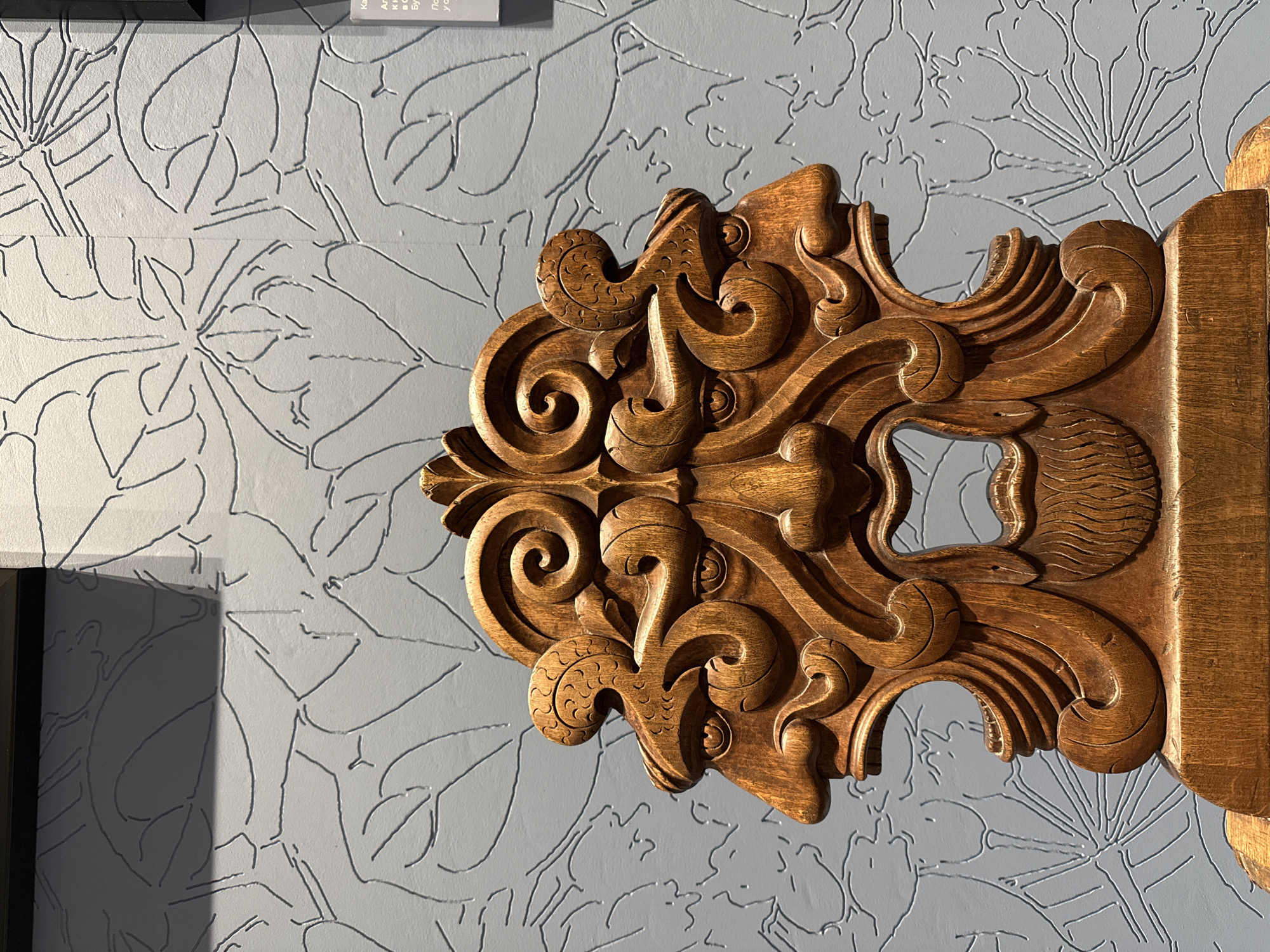A close-up shot shows an intricately carved wooden decorative element mounted on a light gray wall. The wooden piece is stained a warm brown, with detailed swirls and flourishes. The wall behind it has a repeating floral pattern outlined in a darker gray, creating a textured background. A framed sign with text is visible at the top of the image, only partially in the frame. The subject matter is likely a detail found in a building in the city of Perm, Russia.