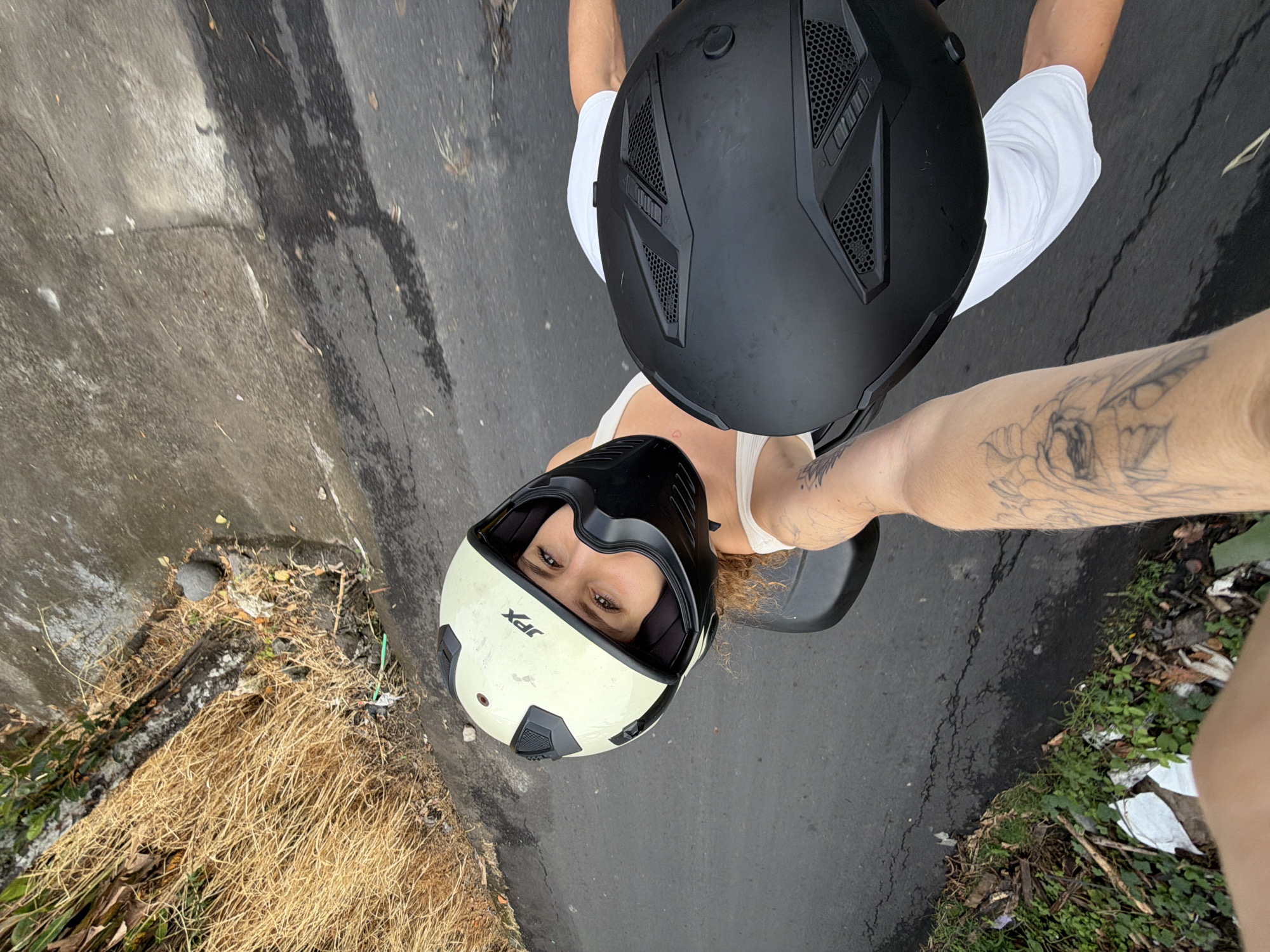This is a first-person point-of-view image taken on a paved road in North Kuta, Indonesia. The foreground shows a person's tattooed arm extending forward, holding a black motorcycle helmet. The helmet is large and covers most of the right side of the frame. Partially visible behind the black helmet is a woman wearing a cream-colored motorcycle helmet with a black chin guard and a white tank top. She is looking directly at the camera with a smile. Her curly, light brown hair is visible escaping the helmet. The background consists of a dry, grassy embankment on the left and the paved road surface with some cracks and water stains. A small rock and some debris are visible on the embankment. The overall scene suggests a motorcycle ride in an outdoor, possibly rural or semi-rural, setting.