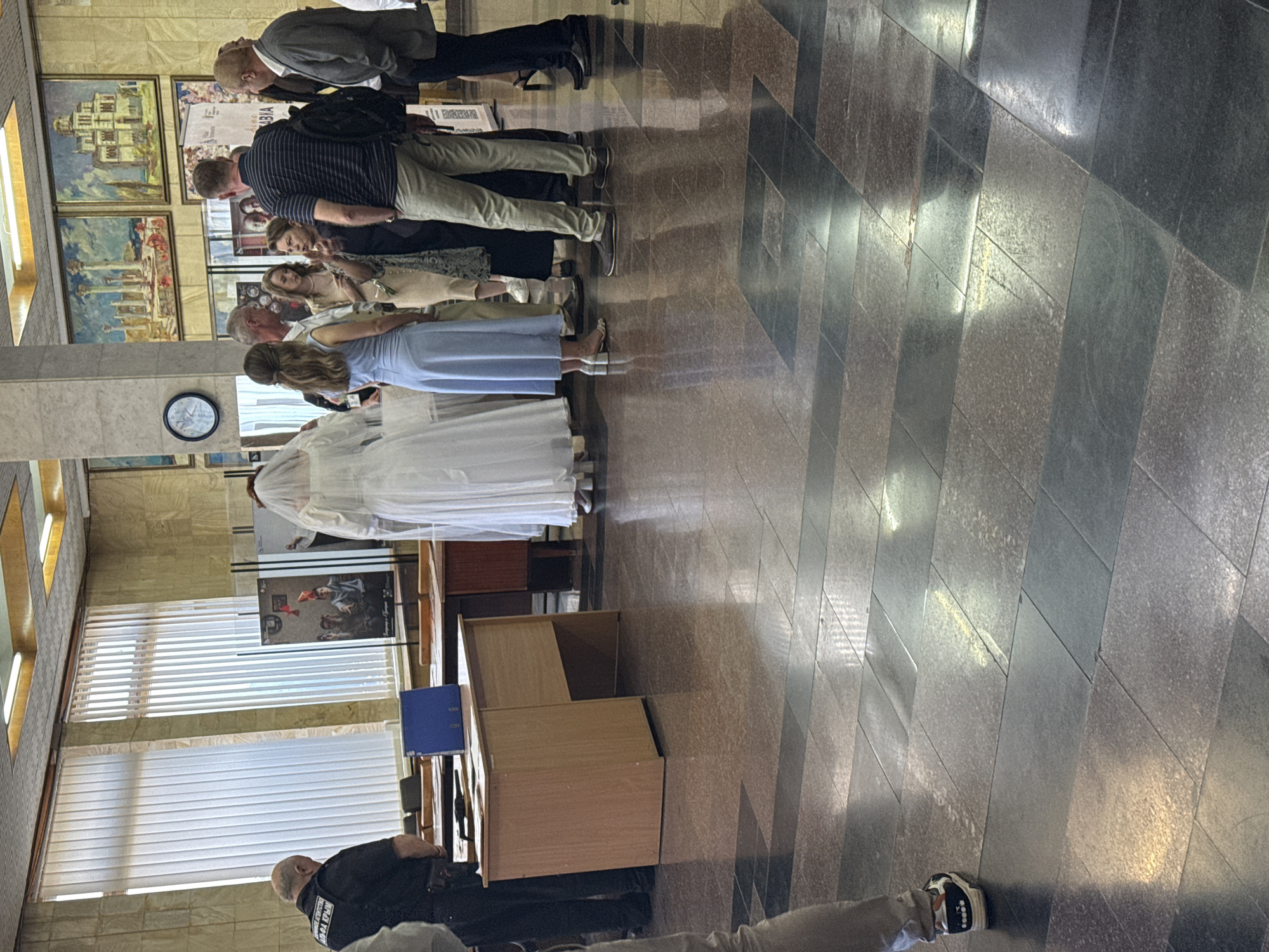 An indoor scene depicts a group of people standing in what appears to be a public building with a polished, tiled floor and high ceilings. The room is furnished with wooden desks and a large blue binder is visible on one of the desks. Paintings adorn the walls, and blinds cover the windows. In the foreground, a person in a black uniform stands facing away from the camera. A woman in a white wedding dress and veil is also present, along with several other individuals in casual attire. Some people are conversing, while others are looking around. The overall atmosphere suggests a gathering, possibly a wedding ceremony or a related event, taking place within an official or administrative setting.