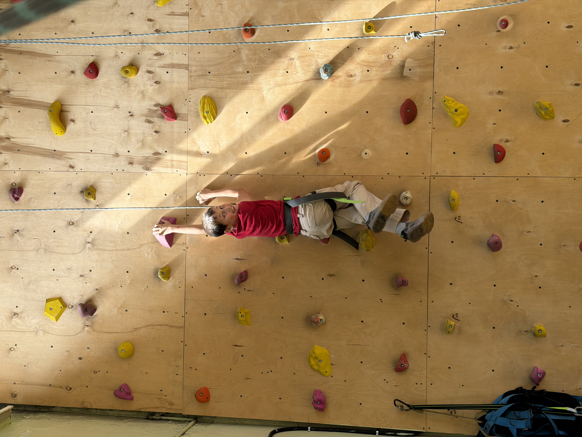A boy is climbing on an indoor climbing wall. The wall is made of wood and has many colorful holds attached to it. The boy is wearing a red shirt, tan pants, climbing shoes, and a harness. He is attached to a rope that runs along the top of the wall. The location is not provided but could be in Izhevsk, Russia.