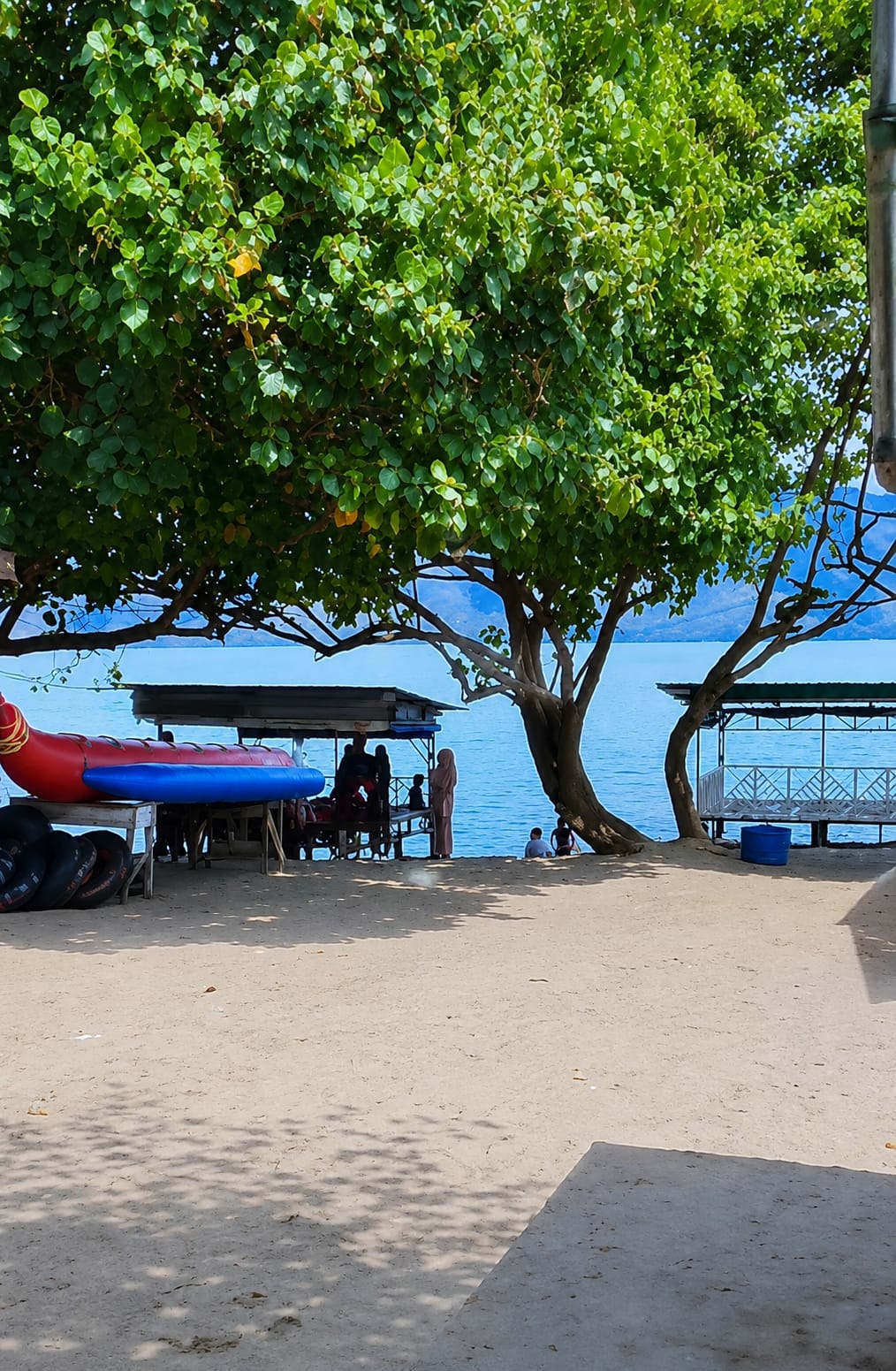 This is a photograph of a beach scene in Situngkir, Indonesia. The foreground is a sandy beach with shadows cast by trees. In the mid-ground, a bright blue body of water is visible, with a distant shoreline of mountains or hills. To the left, a red and blue inflatable boat is resting on a wooden platform alongside several black tires, presumably for water activities. Behind the inflatable boat is a covered structure with a corrugated roof, where several people are gathered. To the right, another covered structure with white railings stands near the water. Two individuals are seated near the water's edge in the right mid-ground, and a person in a light-colored headscarf and dress stands near the covered structure on the left. A large tree with lush green foliage dominates the left side of the frame, its branches extending over the beach and water.