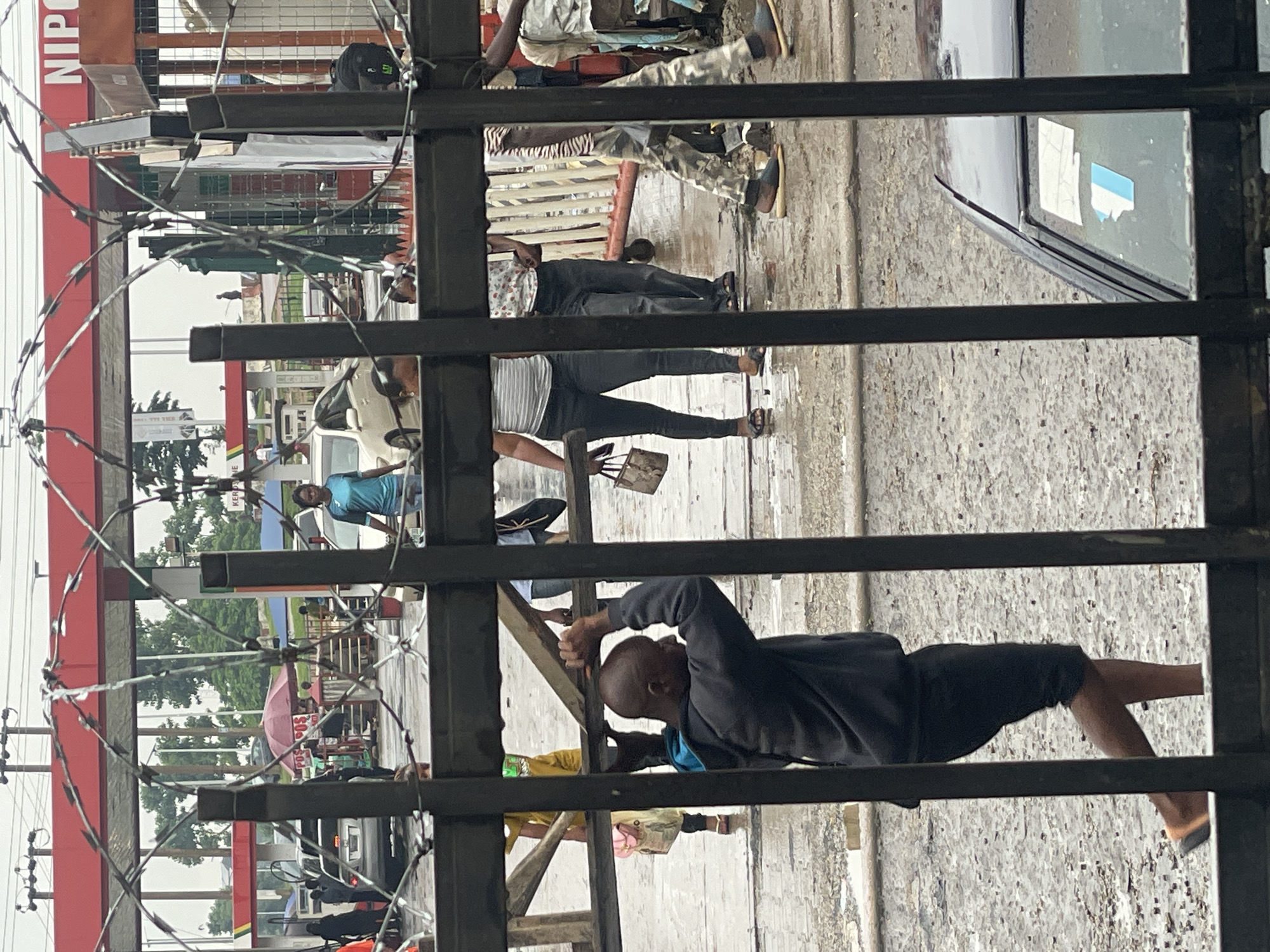 The image depicts a street scene through a metal barrier with barbed wire. In the foreground, a bald Black man wearing a dark hooded sweatshirt and black shorts is holding onto the barrier, looking towards the right. Behind him, several people are walking on a wet, paved surface, some carrying bags. A silver car is parked in the background. In the middle ground, there is a red and white structure, possibly a gas station canopy with a sign that reads "NIPOST". There are other vehicles and people around this structure. The overall environment appears to be an urban street, with trees and overcast skies visible in the distance. The presence of the NIPOST sign suggests the location could be Abuja, Nigeria, as NIPOST is the Nigerian Postal Service.