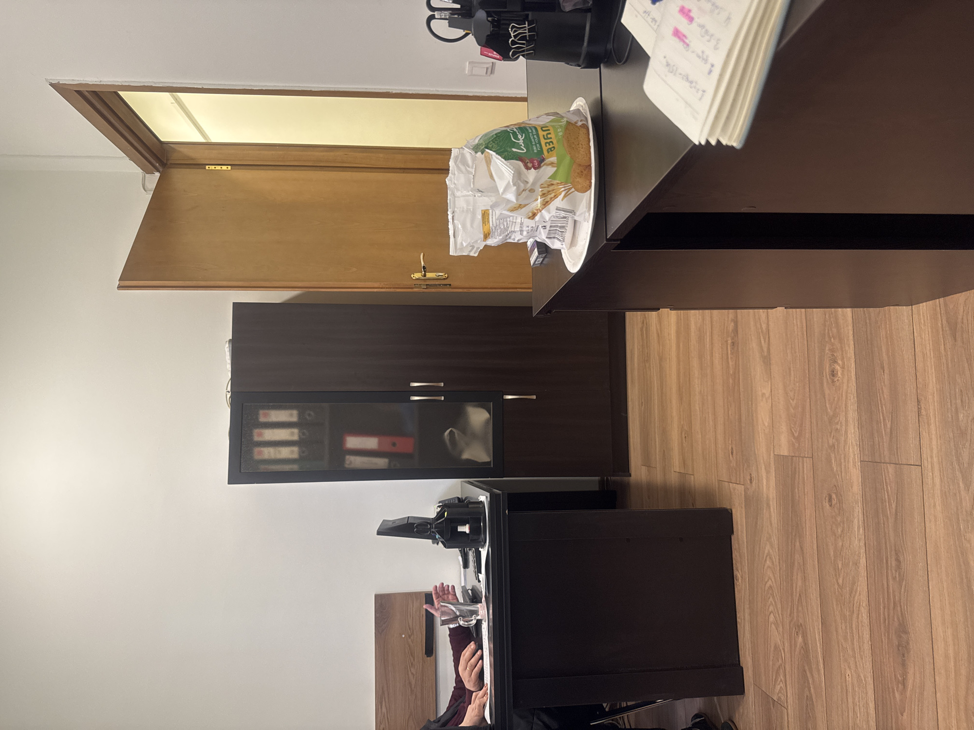 The scene is an interior office setting. On a dark brown desk, there is a black desk lamp and a half-eaten bag of "LUZER" branded snack crackers on a white plate. Next to the snack, there's a notebook with pink annotations on top of the desk. A partly open wooden door is visible in the background. A dark brown cabinet is mounted on the wall below the doorway. A computer monitor sits on another dark brown desk. A person is seated in front of the monitor. The floor is a light brown wood laminate. The location appears to be in an office in Tbilisi, Georgia, based on the brand of snack food.