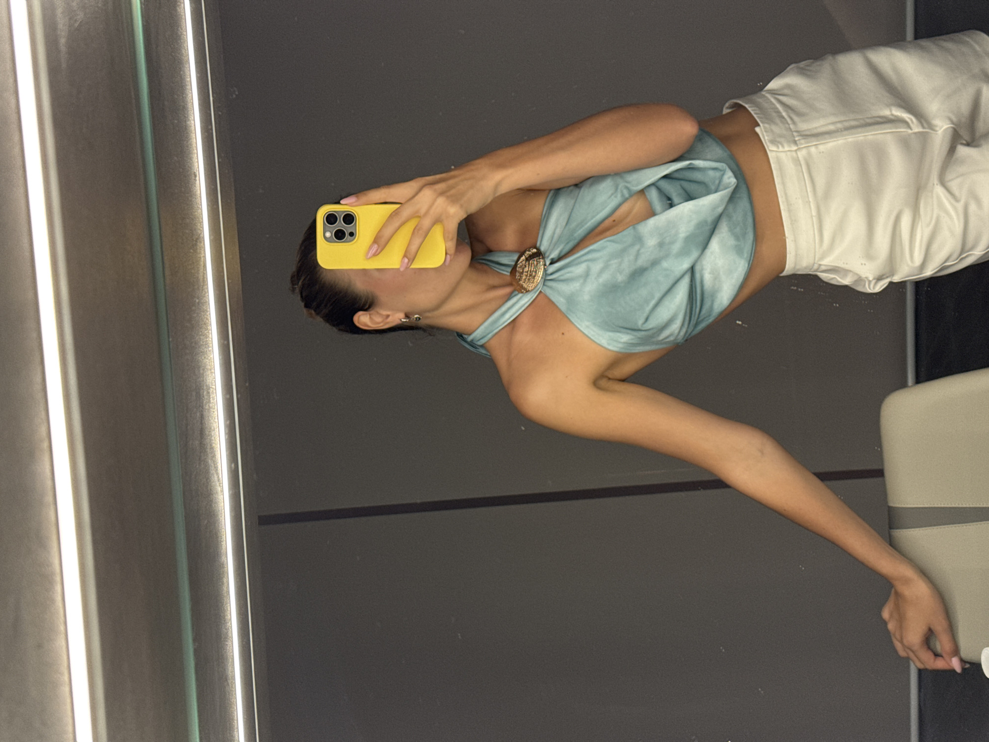 A woman takes a mirror selfie in Unknown, Unknown. She is wearing a light blue halter top secured with a large, round ornament and white pants. Her phone, encased in a yellow case, obscures her face. The scene appears to be inside a modern building or space with grey walls and a reflective mirror. A neutral-colored, possibly beige, object is partially visible on the right side of the image.