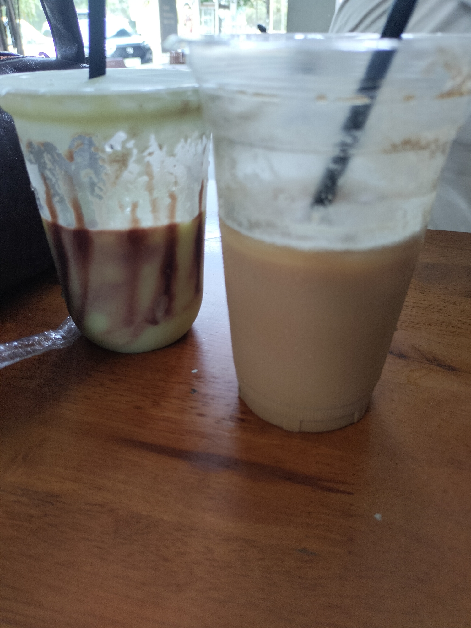 Two plastic cups with straws are on a wooden table. The cup on the left contains a light green beverage with chocolate syrup drizzled down the sides. The cup on the right contains a light brown beverage. The scene is indoors, with a blurred background that includes furniture and possibly a window. The context of Keudealue Ie Puteh, Indonesia is external to the visual information provided in the image.