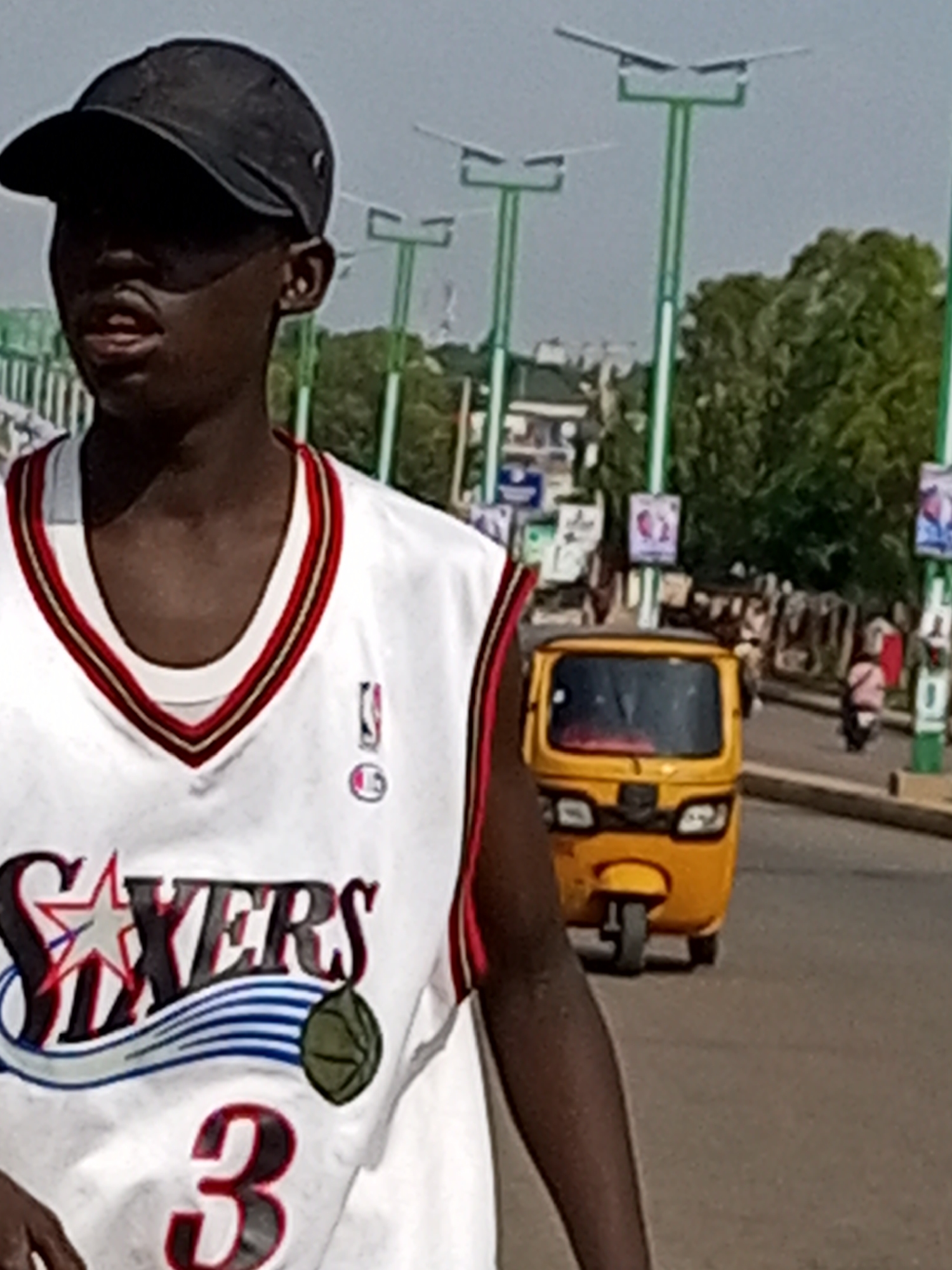The image shows a young Black male wearing a white basketball jersey with the number 3 and "SIXERS" printed on it, along with a black baseball cap. He is standing in the foreground, partially obscuring a yellow auto-rickshaw in the background, which is driving on a paved road. The scene appears to be a street in Tumfure, Nigeria, with streetlights and trees visible. Several other people and vehicles are present in the background, indicating a busy urban environment. The overall lighting suggests it is daytime.