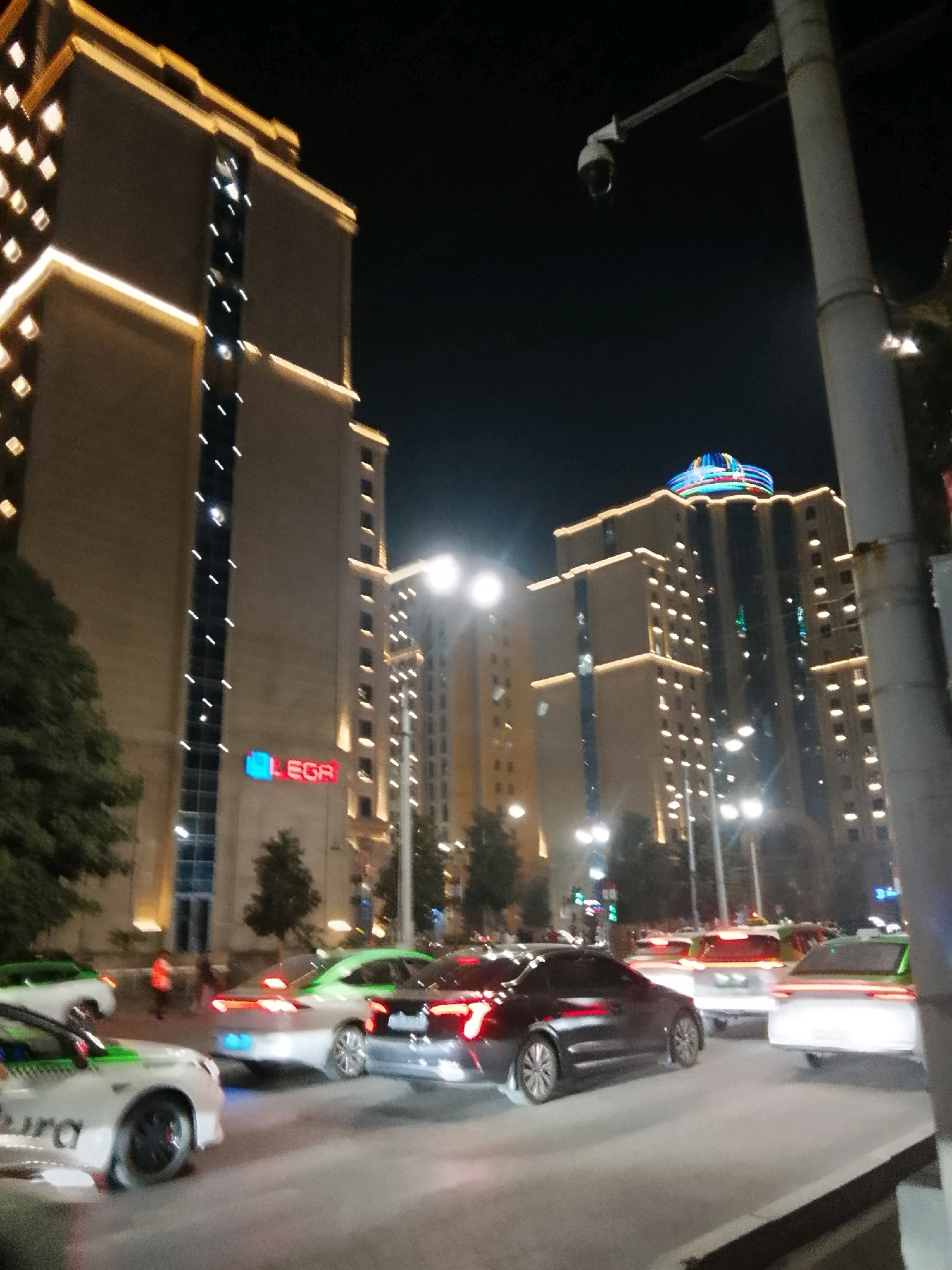 The image shows a nighttime scene in Dushanbe, Tajikistan, featuring modern buildings and traffic on a city street. In the foreground, several cars are visible, including a white car with green accents, a silver car with green accents, and a dark-colored sedan. The cars are on a paved road, suggesting a city environment. Behind the cars, large, illuminated buildings rise into the night sky. One building on the left has a prominent "LEGR" logo in red and blue. Another building in the center is topped with a colorful, circular illuminated structure. Streetlights and lights from the buildings create a bright, urban atmosphere. A security camera is mounted on a pole in the upper right portion of the image. Trees are visible on the left side of the frame. The overall impression is a busy urban intersection at night, illuminated by artificial lights.