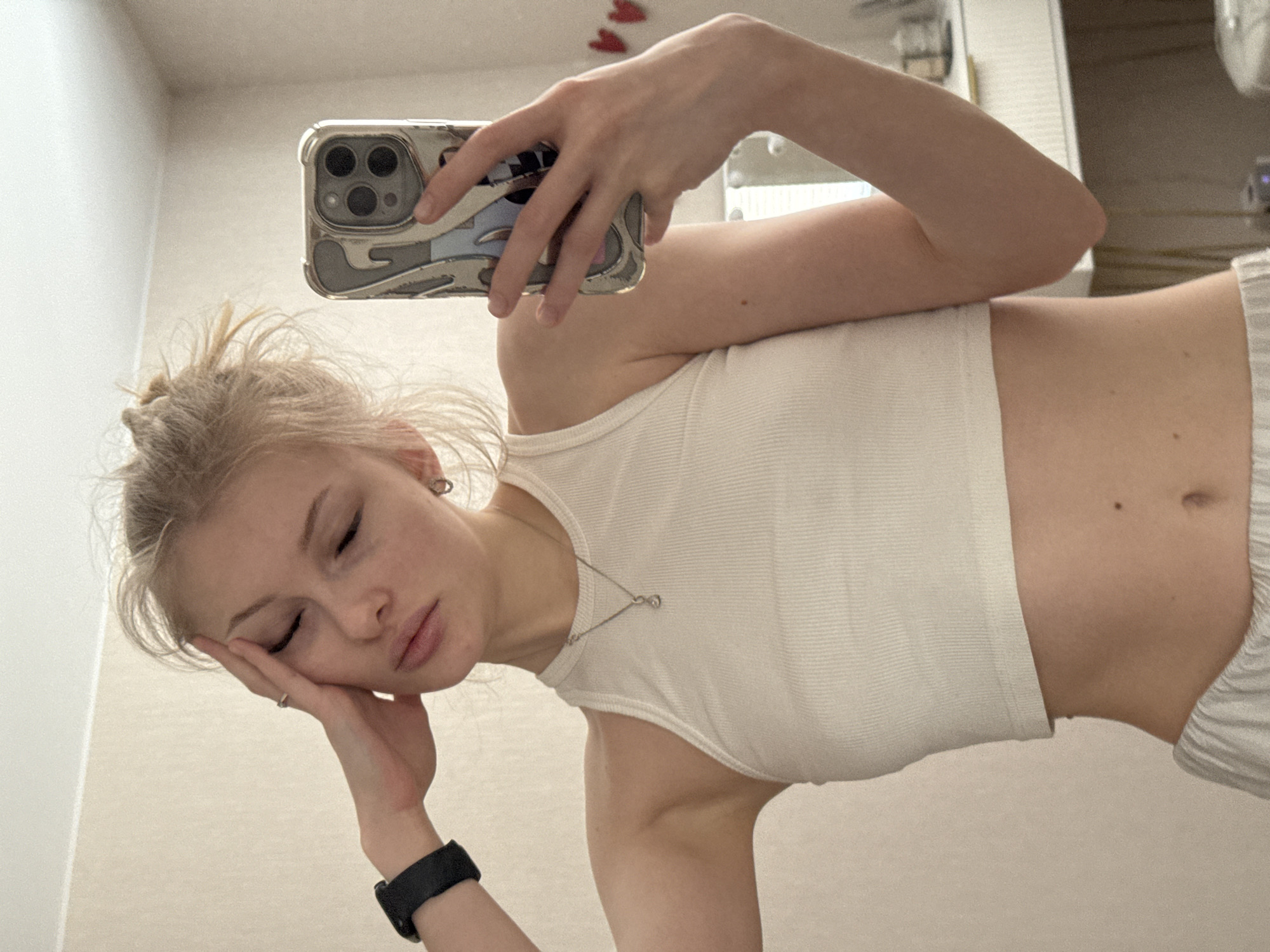 A fair-skinned, blonde woman is taking a selfie in what appears to be an indoor setting, possibly a bathroom. She is wearing a white tank top and light-colored bottoms, with a black smartwatch on her left wrist and a silver necklace. Her eyes are closed, and her head is resting on her hand, suggesting a relaxed or tired pose. The phone case has a light-colored pattern. There's a mirror visible behind her, reflecting a countertop with various items on it. No elements in the photograph conclusively place the location in Perm, Russia.