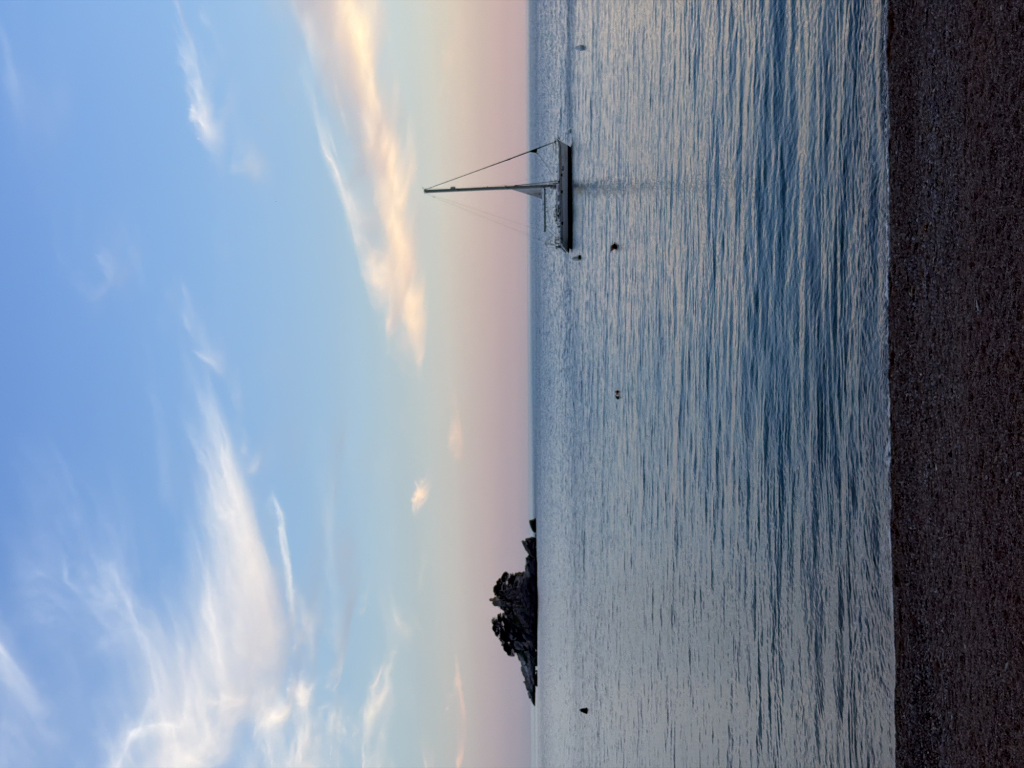 The image depicts a sailboat anchored in the sea off the coast of Petrovac centar, Montenegro. The scene captures the transition between sea and sky during either sunrise or sunset. The sky features hues of blue, pink, and gold, with wispy clouds. A small, rocky islet is visible in the distance. The sea reflects the colors of the sky and has gentle ripples on the surface. The shoreline on the right side of the image is covered with dark pebbles.