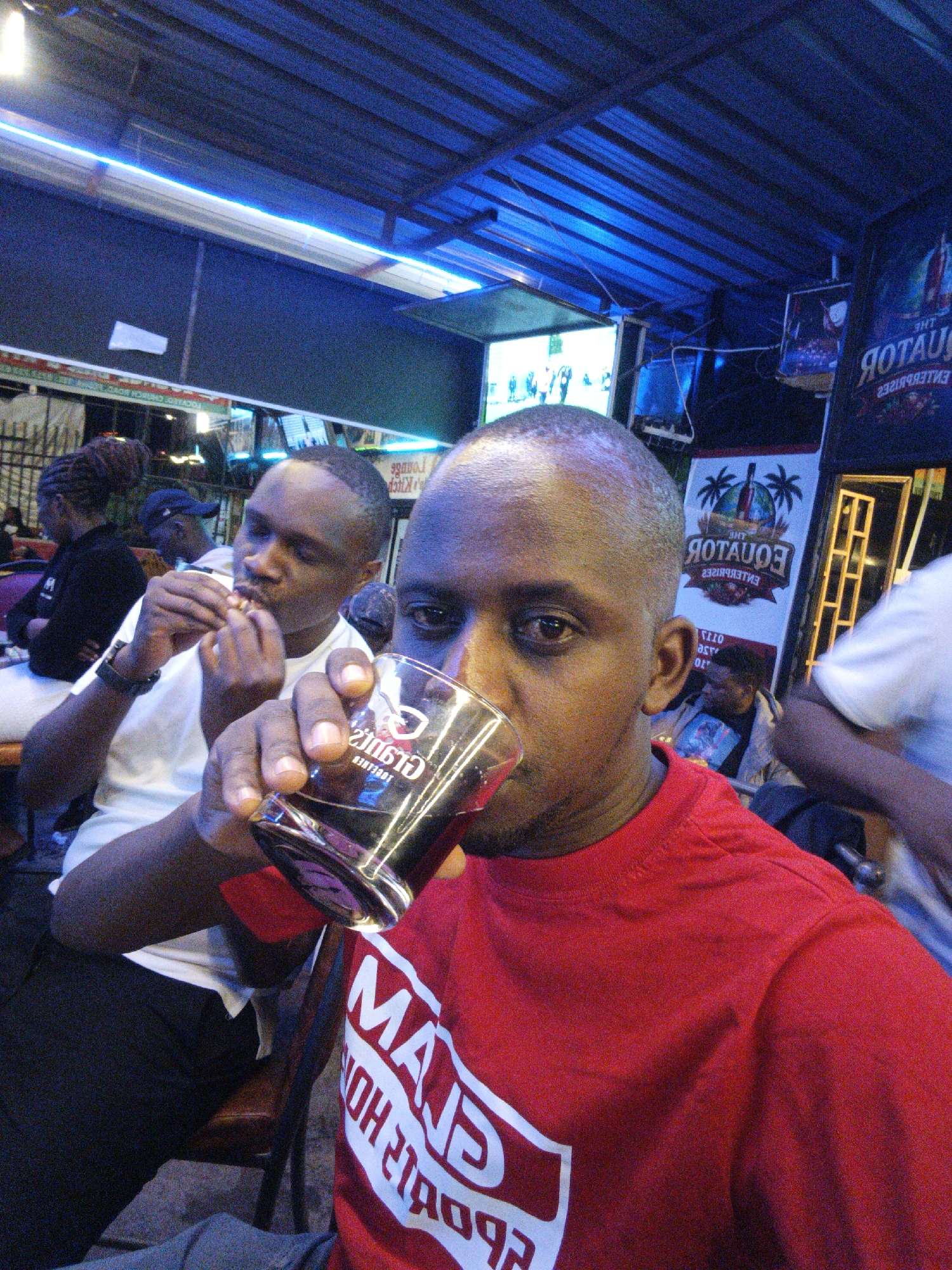 The image shows two men at what appears to be an outdoor bar. The man in the foreground is wearing a red t-shirt and is drinking from a glass. The other man is wearing a white t-shirt and appears to be eating something. There are other people in the background, as well as a television screen. A banner says "The Equator Enterprises", indicating the location is likely Nairobi, Kenya.