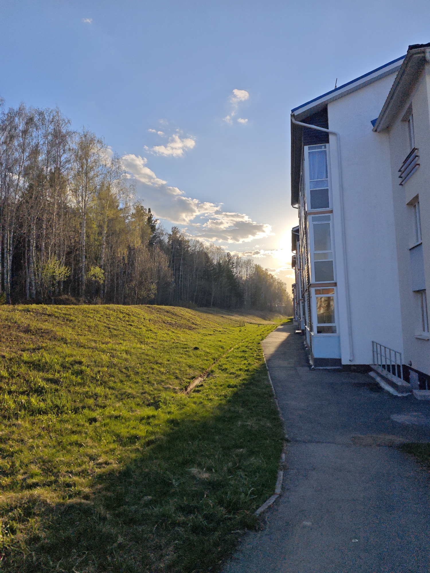 The image shows a view in Мичуринский, Russia, featuring a multi-story white building to the right, partially obscuring the view of the setting sun. The building has blue trim and several vertical window bays. A paved walkway runs along the side of the building. To the left of the walkway is a grassy embankment, and beyond that, a line of trees with some new spring foliage. The sky is blue with scattered clouds, illuminated by the sun. The light suggests it is either sunrise or sunset. No people are visible in the image.