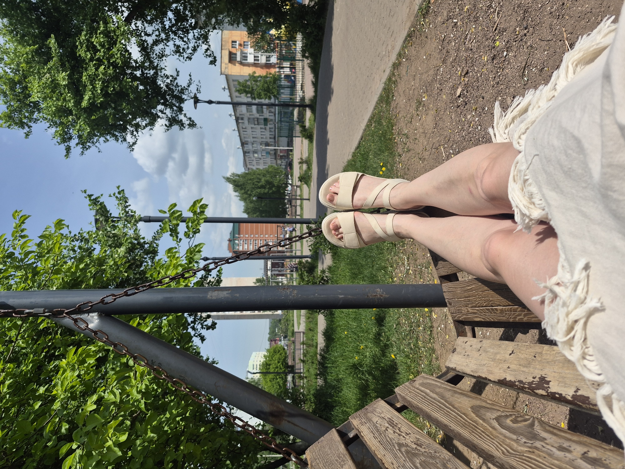 A person is sitting on a wooden park bench with their legs extended, wearing light-colored strappy sandals. The bench is suspended by chains from a dark metal frame. The background shows a city park with green trees, grass, and buildings. A paved path is visible in the background. The weather appears sunny.