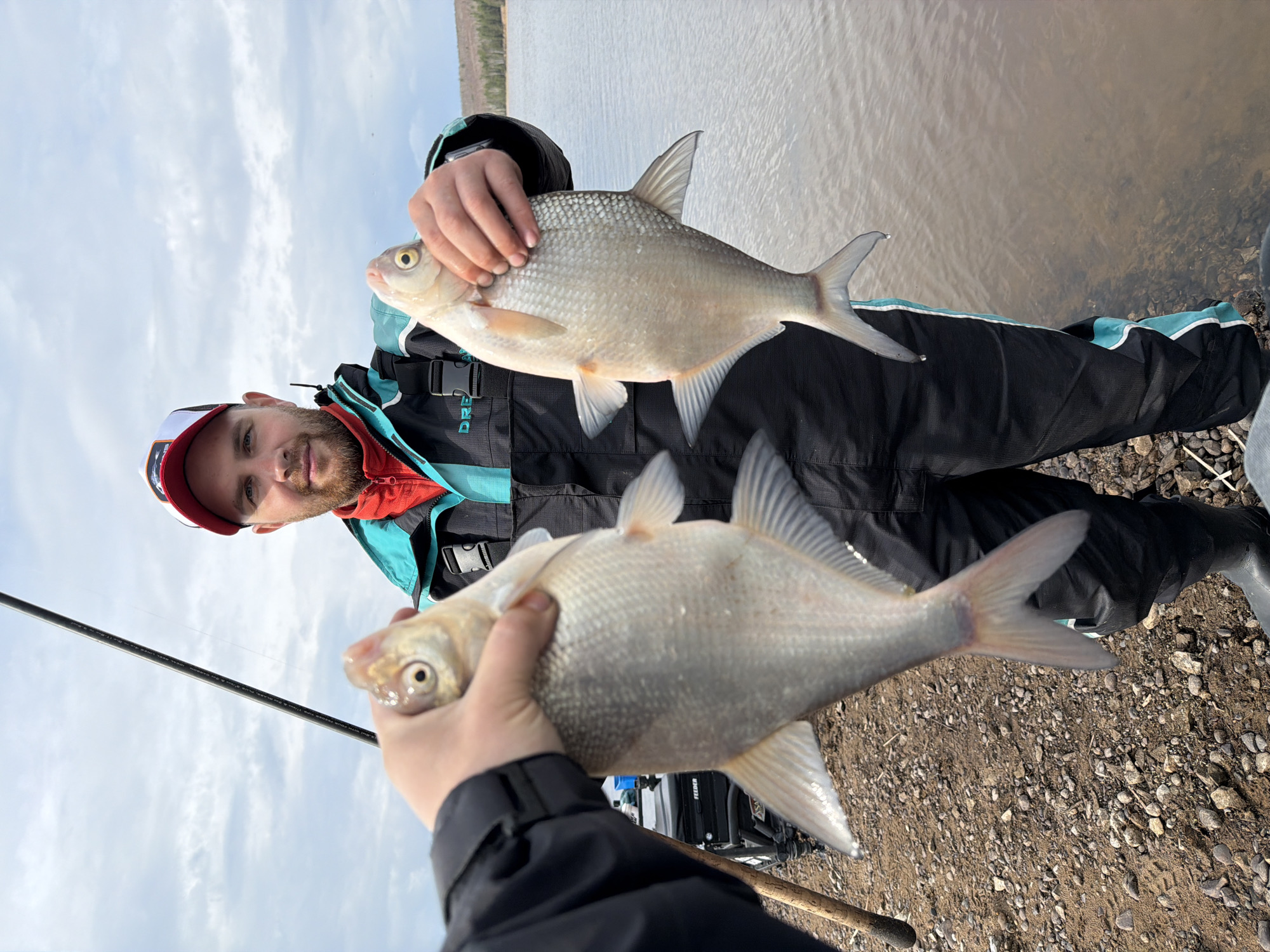A man is holding two fish near the bank of a body of water. The man wears a red and white hat, a red shirt, and a black jacket with teal accents. He is looking at the camera. He is standing near the water's edge, with a gravelly shoreline visible. A fishing rod is visible in the foreground. The photo was taken near the city of Chaikovsky, Russia.