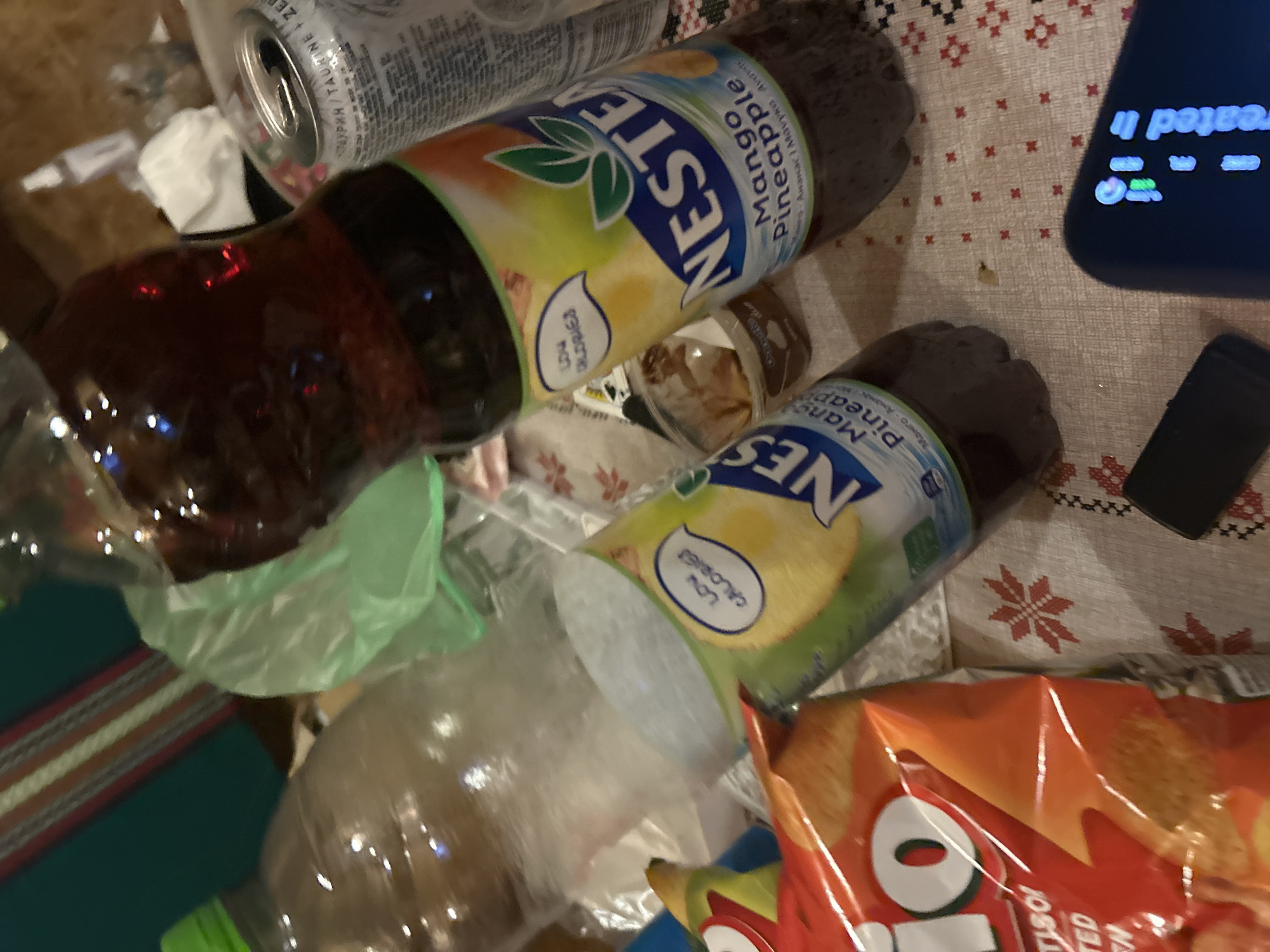 The image shows a close-up of a table with various food and drink items, suggesting a casual gathering or a snack. The scene is indoors, likely a domestic setting given the presence of a patterned tablecloth with red and white embroidered designs, reminiscent of a Christmas or folk theme.
Dominating the foreground are two bottles of "Nestea Mango Pineapple" flavored iced tea. One bottle is in focus on the right, displaying its label clearly, which highlights "LOW CALORIES" and the brand's logo with green leaves. The drink inside is a dark amber color. A second bottle of the same beverage is partially visible on the left. Next to the bottles, a bag of "Rio" brand potato chips is present, its bright orange packaging and the "O" of the logo clearly identifiable.
In the background, a large, clear plastic bottle, possibly containing water, is visible on the left. To its right, a crumpled translucent green plastic bag is partially obscuring other items. A silver can, likely a beverage, is also visible to the right of the focused Nestea bottle, with some text in a foreign language, possibly Cyrillic. A small, dark brown plastic container with a lid, containing a substance that appears to be a dip or dessert, is situated between the two Nestea bottles.
The overall lighting is dim, with some reflections visible on the bottles, suggesting artificial light sources. A dark, rectangular object, possibly a remote control or a small electronic device, lies on the tablecloth in the lower right quadrant of the image, near a dark-colored smartphone screen that displays some illuminated text and icons. The tablecloth itself has a textured, slightly sparkly appearance. The environment is cluttered but organized, with items arranged on the surface of the table. No individuals are present in the frame.