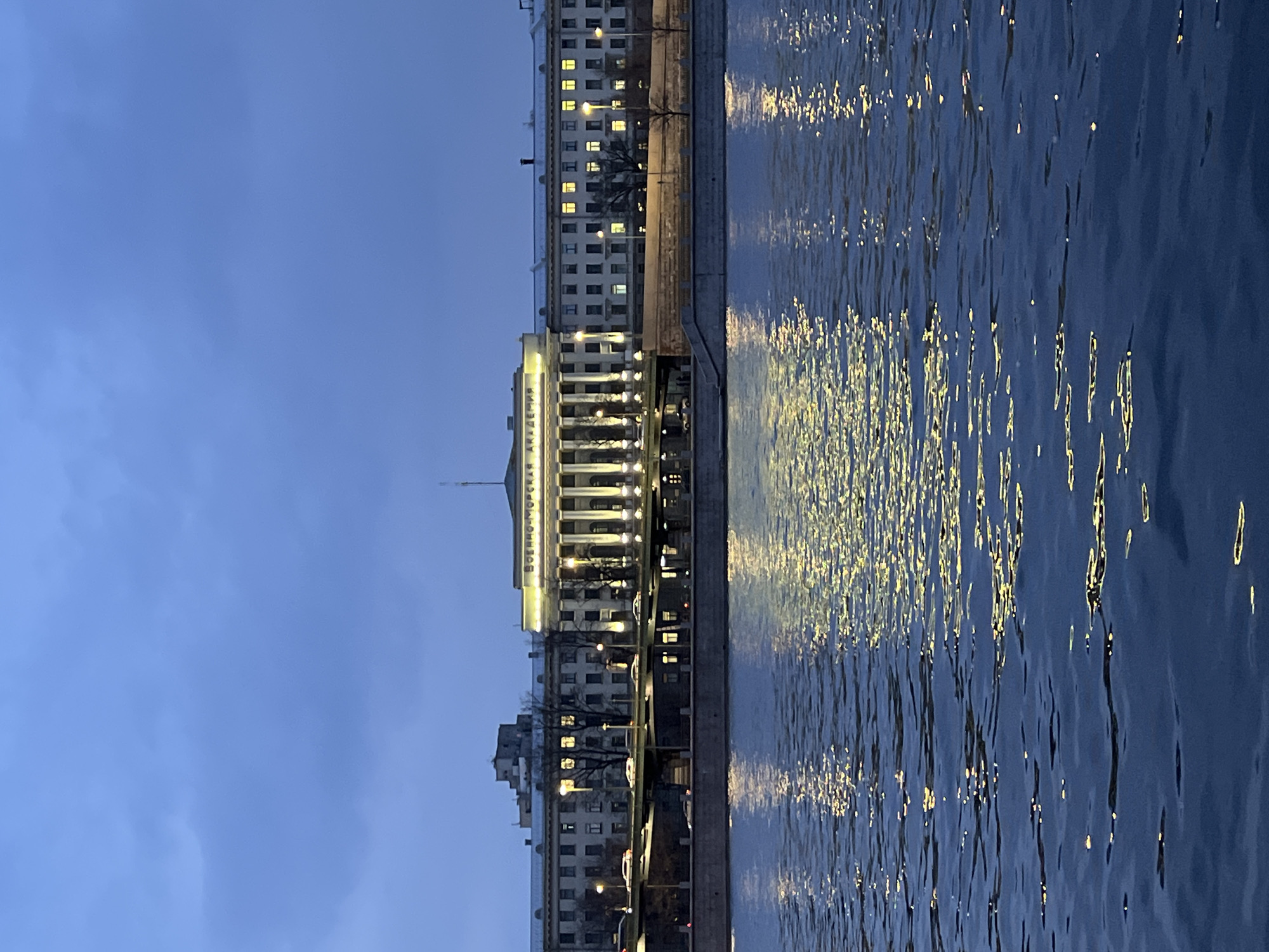 This image captures a nighttime or late dusk scene across a body of water towards an illuminated building complex in Saint Petersburg, Russia. The upper quarter of the frame displays an overcast, dark blue sky. The lower two-thirds of the image consists of rippled, dark blue water reflecting numerous elongated, bright yellow-white light streaks originating from the opposite bank.
Above the water, a grey concrete embankment supports a multi-lane road featuring multiple streetlights emitting yellow-white light. Several light-colored passenger vehicles, some exhibiting motion blur, are visible on this road. Sparse, leafless trees line the embankment.
The dominant subject is a long, multi-story building complex. Its central portion presents a classical facade with a prominent colonnade, brightly illuminated by yellow-white lights. Above the columns, the Cyrillic inscription "ВОЕННО-МОРСКАЯ АКАДЕМИЯ" (Naval Academy) is clearly visible. A flagpole stands on the roof directly above this central section. Flanking the central edifice are less ornate building sections, each with multiple windows, many of which are illuminated from within. This structure is identified as the Naval Academy, situated along a waterfront in Saint Petersburg.