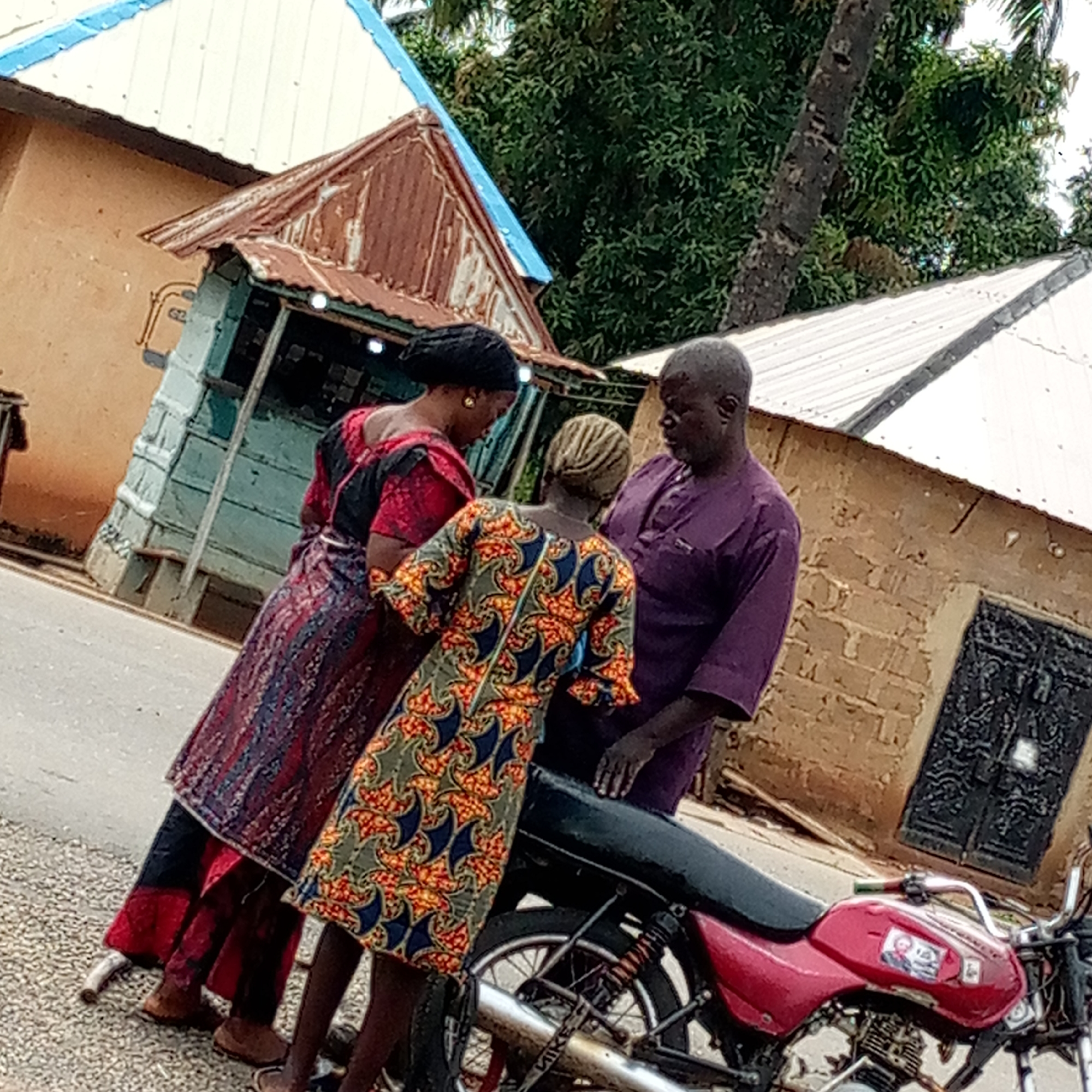 In Mararaba, Nigeria, three people stand by a parked red motorcycle on an asphalt road next to a small, blue-painted, corrugated-roofed shop. A woman on the left, wearing a long, dark red and black patterned dress, is turned towards the others. The person in the middle, also a woman, wears a dress with orange, blue, and green patterns. A man in a purple short-sleeved shirt stands on the right, facing the two women. Behind them are buildings with corrugated iron roofs; one building has a tan-colored facade and a black, ornate door. A tree is visible in the background.