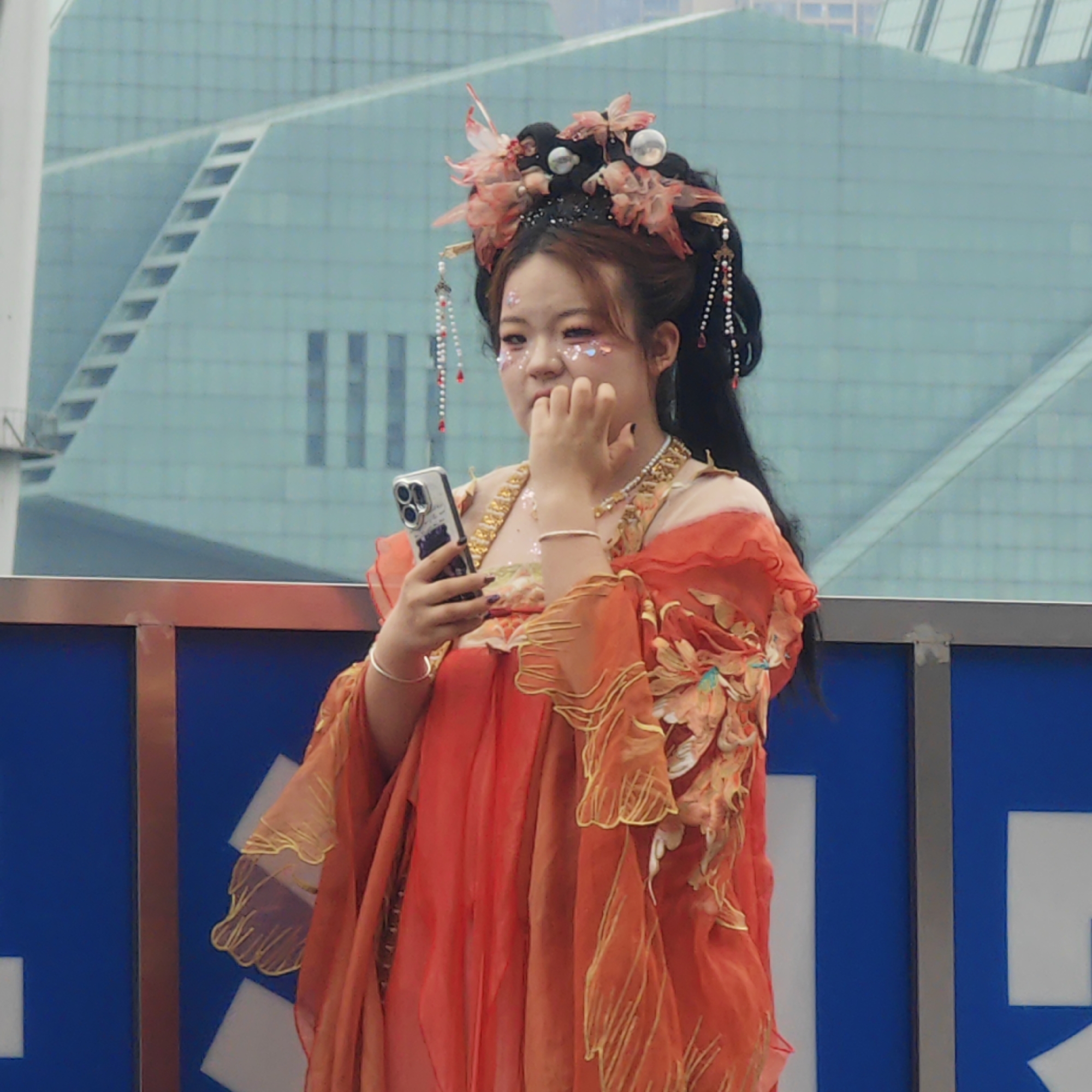 A medium shot shows a young woman in traditional Chinese dress holding a smartphone. She has decorated her face with glittery stickers. Her hair is arranged with flowers and beads. She is standing behind a blue and silver railing, with a modern glass building in the background. The location is Yuzhong District, China.