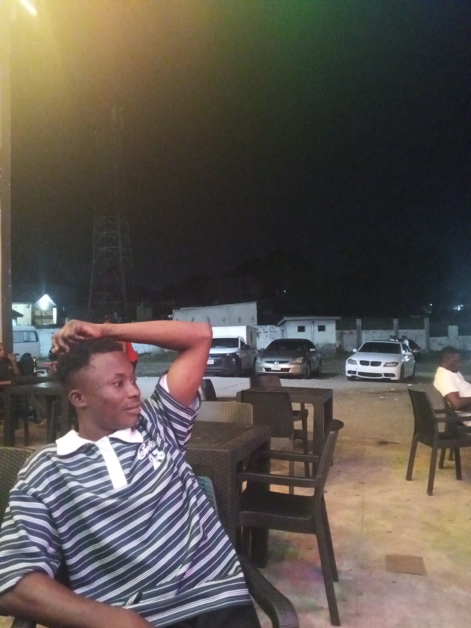 The image captures an outdoor night scene, likely a casual gathering place such as a restaurant or bar. In the foreground, a young man of African descent is seated on the left. He is wearing a short-sleeved polo shirt with horizontal white and dark blue/black stripes and a white collar. His left arm is bent, with his hand resting on the back of his head or neck, indicating a relaxed posture, and he is looking towards the right side of the frame.
The setting includes dark-colored, possibly wicker, tables and chairs arranged around the seating area. The ground surface is light-colored, showing subtle, colorful reflections from ambient lights. In the mid-ground and background, there is a parking area with several cars, including a distinct white BMW sedan on the right and another light-colored sedan. Beyond the vehicles, low-rise buildings and a tall metal power pylon are visible against the dark night sky. Another person, wearing a white t-shirt, is partially visible seated on the far right.
The time of day is clearly night, as indicated by the dark sky and the reliance on artificial lighting, which casts a warm glow from sources outside the frame and illuminates the scene. The overall atmosphere appears calm and social. There is some faint, unreadable text or a logo on the sleeve of the main subject's striped shirt.