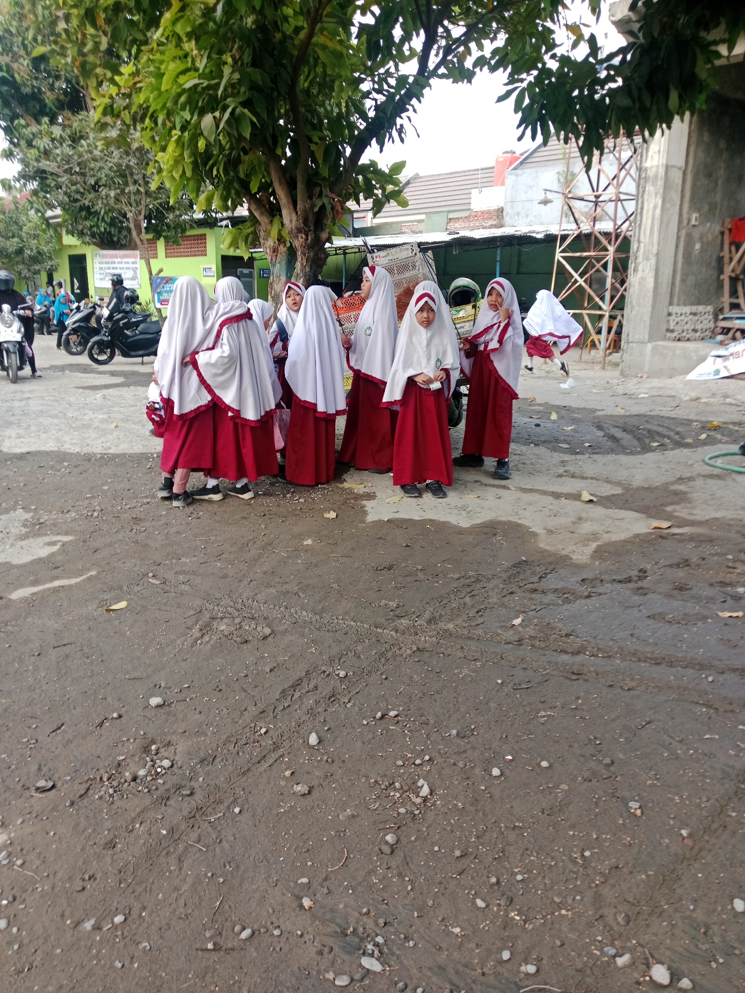 A group of seven young girls, dressed in school uniforms consisting of white hijabs and red skirts, stand in a dirt lot. The girls are gathered around a tree with green leaves, and some are facing away from the camera. In the background, there are buildings, a construction site with scaffolding, motorcycles, and other students. The ground is covered in dirt, pebbles, and some fallen leaves. The overall scene appears to be a schoolyard in Kediri City, Indonesia, possibly during dismissal or a break time.