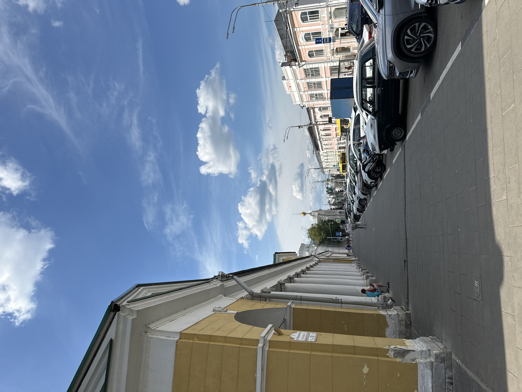A sunny day in Arkhangelsk, Russia, features a street scene with a yellow, colonnaded building on the left and a row of parked cars on the right. The colonnade has arched entrances and is supported by white pillars. A person is sitting on a bench under the colonnade, looking at their phone. Further down the street, a few more people are walking. Across the street, several buildings with pink and white facades line the road, and a golden-domed church is visible in the distance. The sky is blue with scattered white clouds. The street is paved with gray tiles, and white lines mark the parking spaces. A street lamp is visible on the right side of the image.