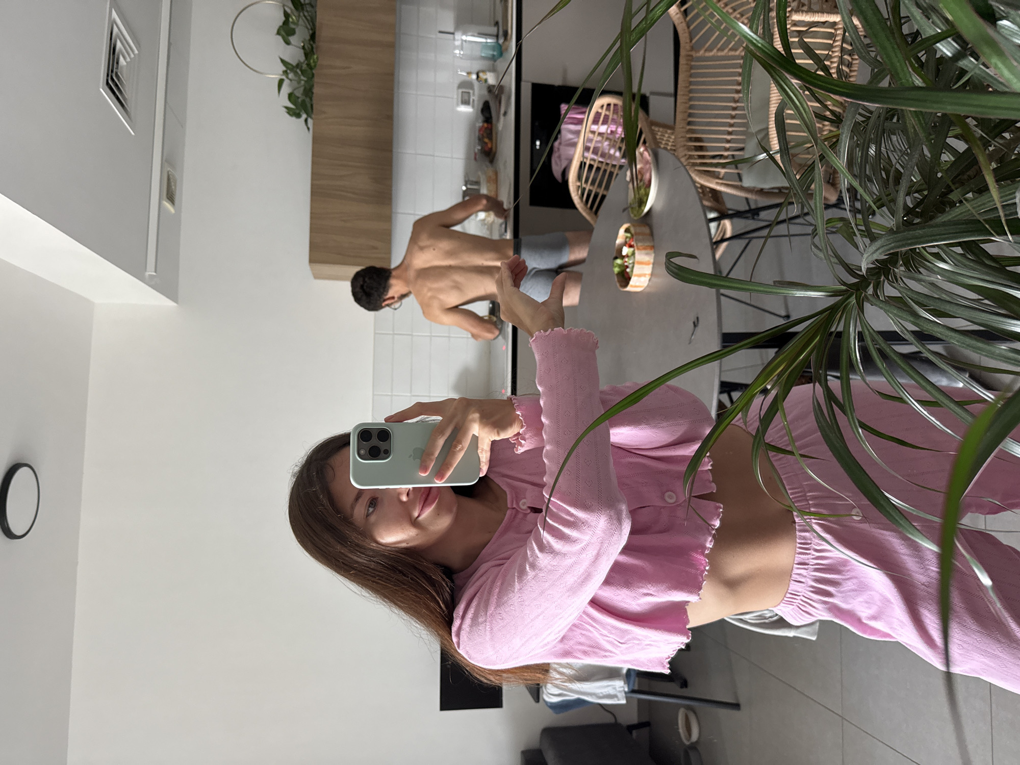 A mirror selfie shows a young woman in a pink top and pants. She is holding a phone to take the picture. In the reflection, a shirtless man is standing at a kitchen counter. The kitchen is white and minimalist. There's a round dining table with chairs nearby and a plant in the foreground, slightly obscuring the view. The location is likely an apartment in Tel-Aviv, Israel, based on the typical architectural style and interior design of residential spaces in the city.