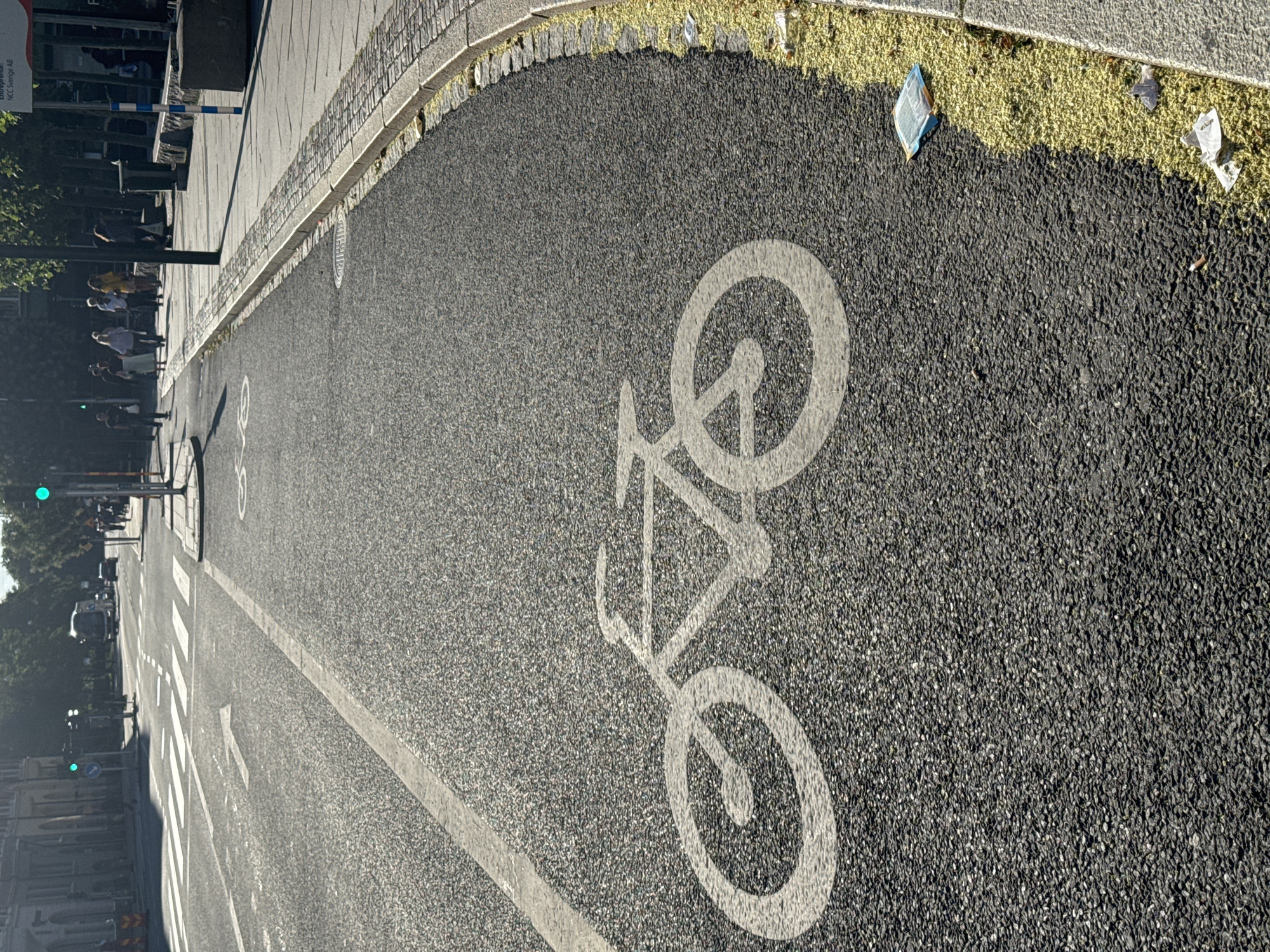 A street scene with a bike lane painted on asphalt. A white stencil of a bicycle is visible in the bike lane. In the background, there is a crosswalk, traffic lights, pedestrians, a bus, and a building. The scene appears to be in an urban environment during the day.