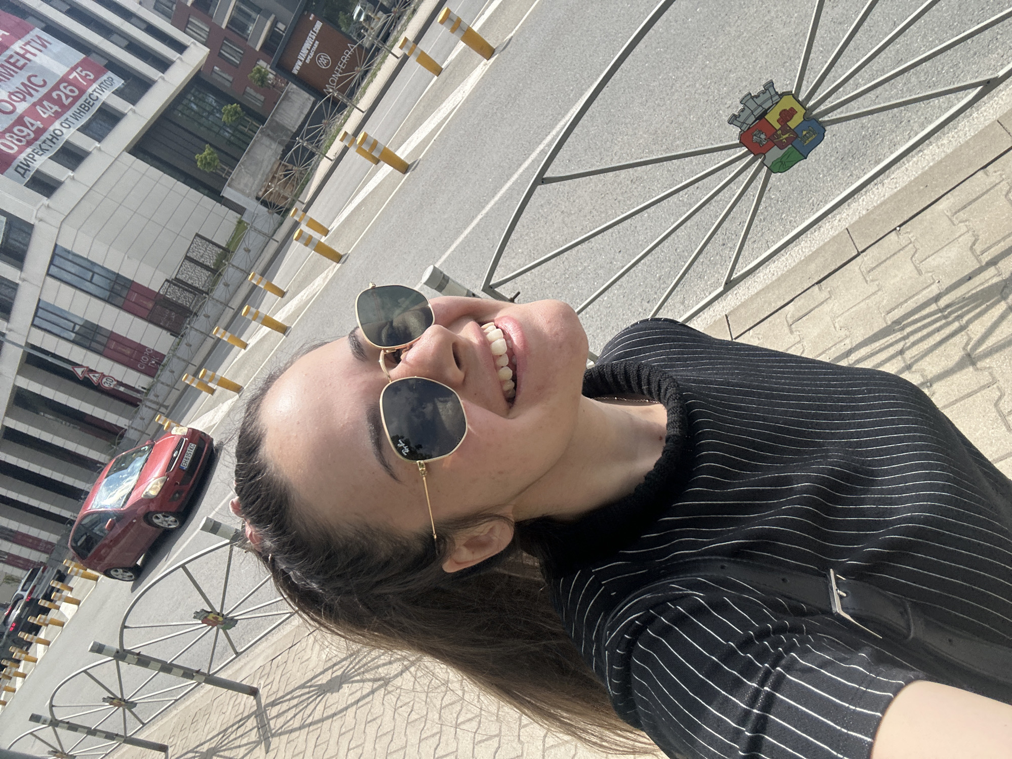 A young woman, seen from a low angle, takes a selfie on a sunny day in an urban setting identified as Sofia, Bulgaria. She is smiling broadly with her mouth open, looking upwards towards the camera or sky, and wearing sunglasses with gold frames (partially revealing "Ray-B" on the lens) and a black and white striped top. A black bag strap is visible over her shoulder.
The background reveals a city street with modern multi-story buildings. One building features large red and white banners with Bulgarian text: "АПАРТАМЕНТИ" (Apartments), "ОФИСИ" (Offices), a phone number "0894 44 26 75", and "ДИРЕКТНО ОТ ИНВЕСТИТОР" (Directly from investor). Another building has a brown sign with "WWW.VAN...VEST.COM" and "MONTERRA".
The street features a paved sidewalk and asphalt road, lined with yellow and white bollards and unique metal fence barriers designed in a spoke-wheel pattern. One of these barriers prominently displays a colorful coat of arms, which is identifiable as the municipal emblem of Sofia, featuring a fortress, a lion, and other symbolic elements. A red Ford Focus car with a Bulgarian license plate "EH 8511 KC" is parked nearby, and a "50" speed limit sign is visible in the distance. The bright light and distinct shadows indicate clear, sunny weather, likely during the day. The woman's joyful expression conveys a sense of happiness and relaxation.