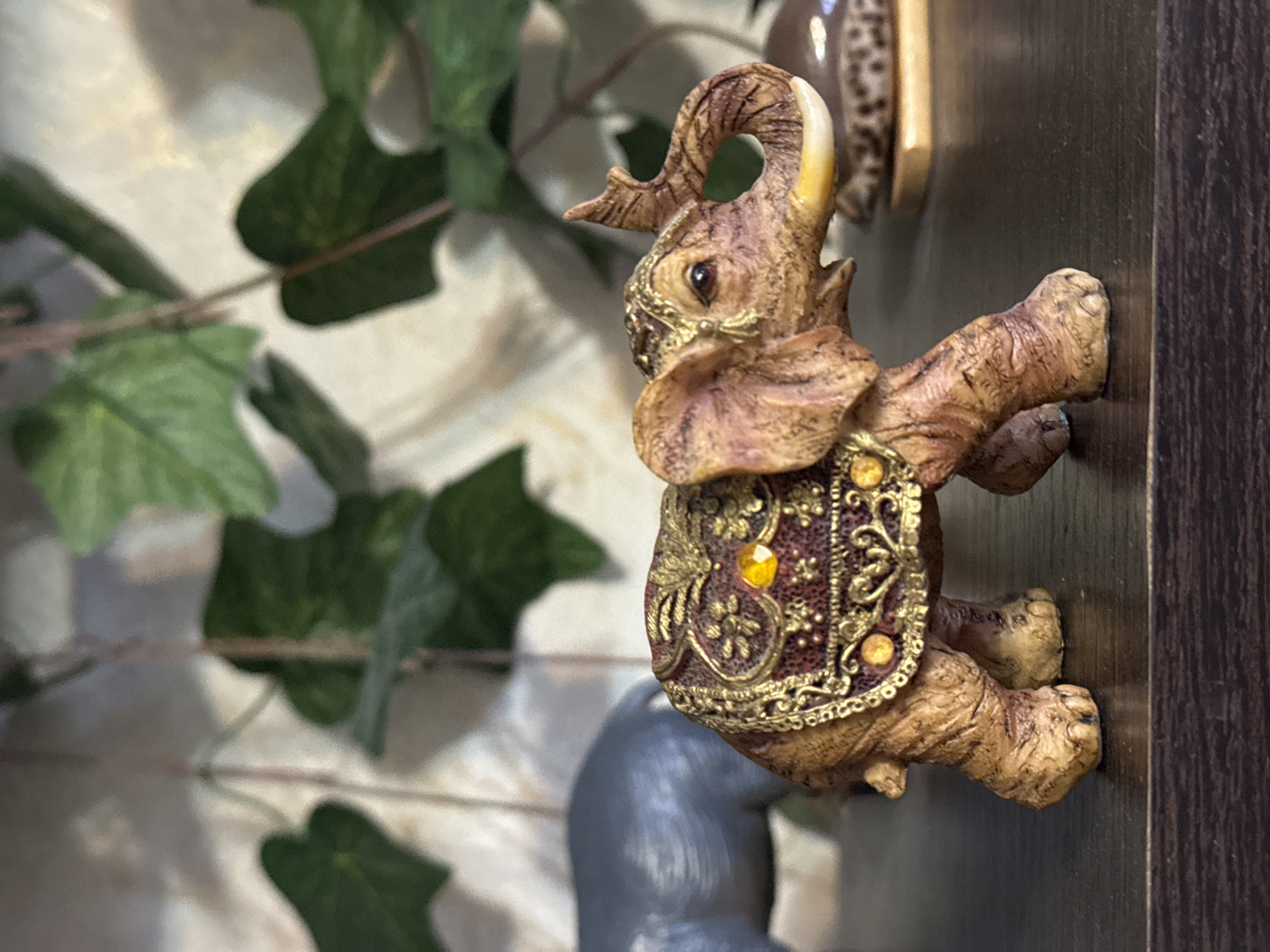 The image shows a decorative elephant figurine, brown in color and adorned with gold embellishments, placed against a backdrop of artificial ivy and a dark wooden surface. The elephant is standing on its hind legs with its front legs extended. The location of the city of Kladovitsy, Russia is not evident in the image.