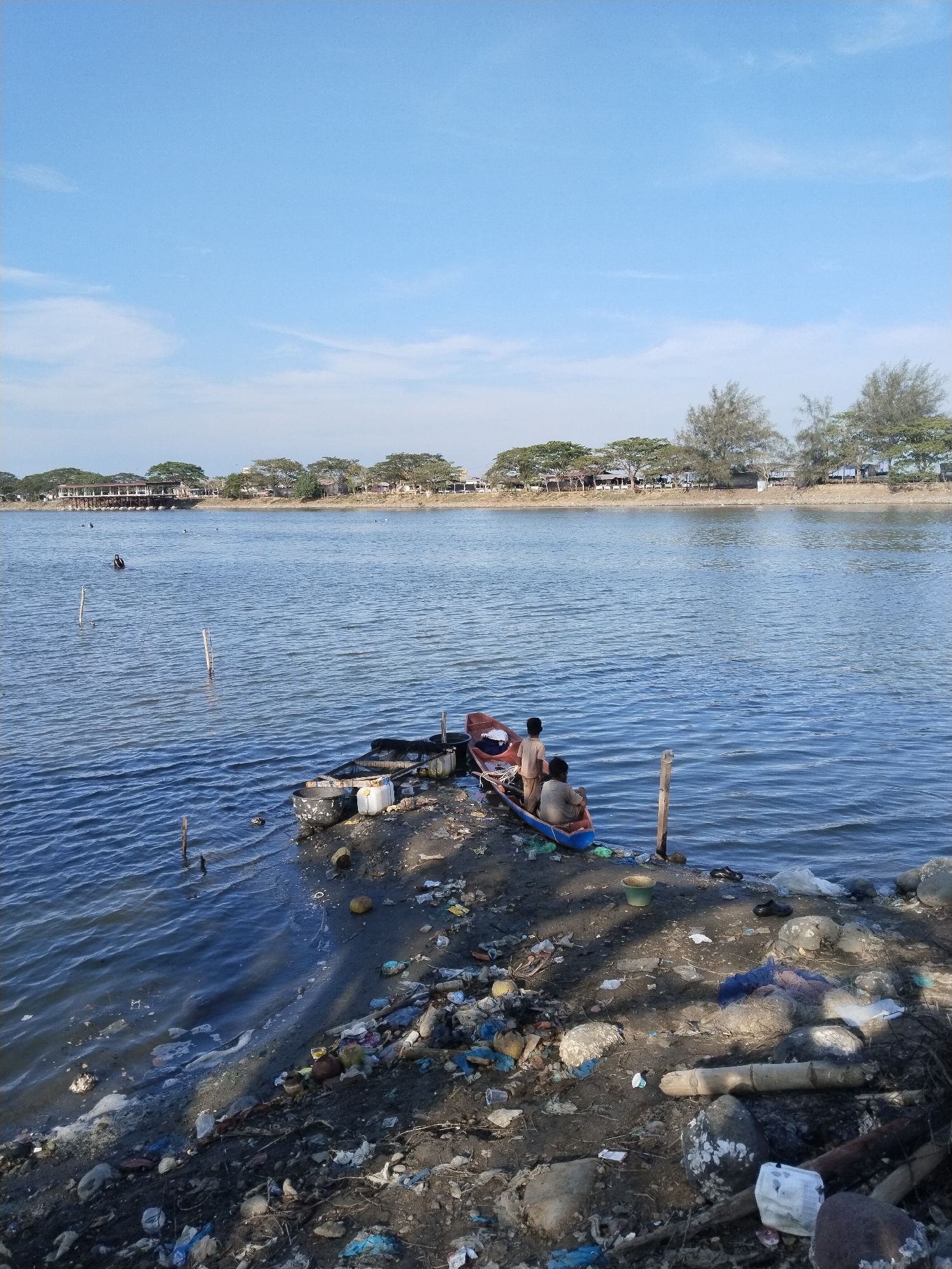 The image captures a wide view of a water body, likely a river or an estuary, under a clear blue sky with a few wispy clouds. The setting appears to be a coastal or riverine area in Lhokseumawe, Indonesia.
In the foreground, a muddy and heavily littered bank stretches along the water's edge. Numerous pieces of trash, including plastic bottles, bags, Styrofoam, fishing nets, coconut husks, and other debris, are scattered across the dark soil. Two men are seated next to a small, narrow boat painted blue and red, which is pulled up onto this cluttered bank. One man, wearing a light-colored shirt, is positioned towards the stern of the boat, facing away from the viewer, while the other, also in a light shirt, is seated within the boat, looking out at the water. They appear to be resting or preparing for an activity. Near the boat, a white plastic jerrycan and a black bucket are visible, amidst more refuse. Several wooden poles are driven into the shallow water close to the shore.
Further out on the water, several dark shapes, possibly people or objects, can be seen, with at least one distinct figure appearing to be swimming or wading. The distant bank features a line of green trees and various buildings, including some with reddish roofs, suggesting a community or settlement beyond the waterway.
The time of day is bright, likely mid-morning or afternoon, indicated by the clear sunshine and visible shadows. The overall weather is calm and sunny. No visible text is present in the image. The scene presents a juxtaposition of daily life and natural beauty with significant environmental pollution along the immediate shoreline.