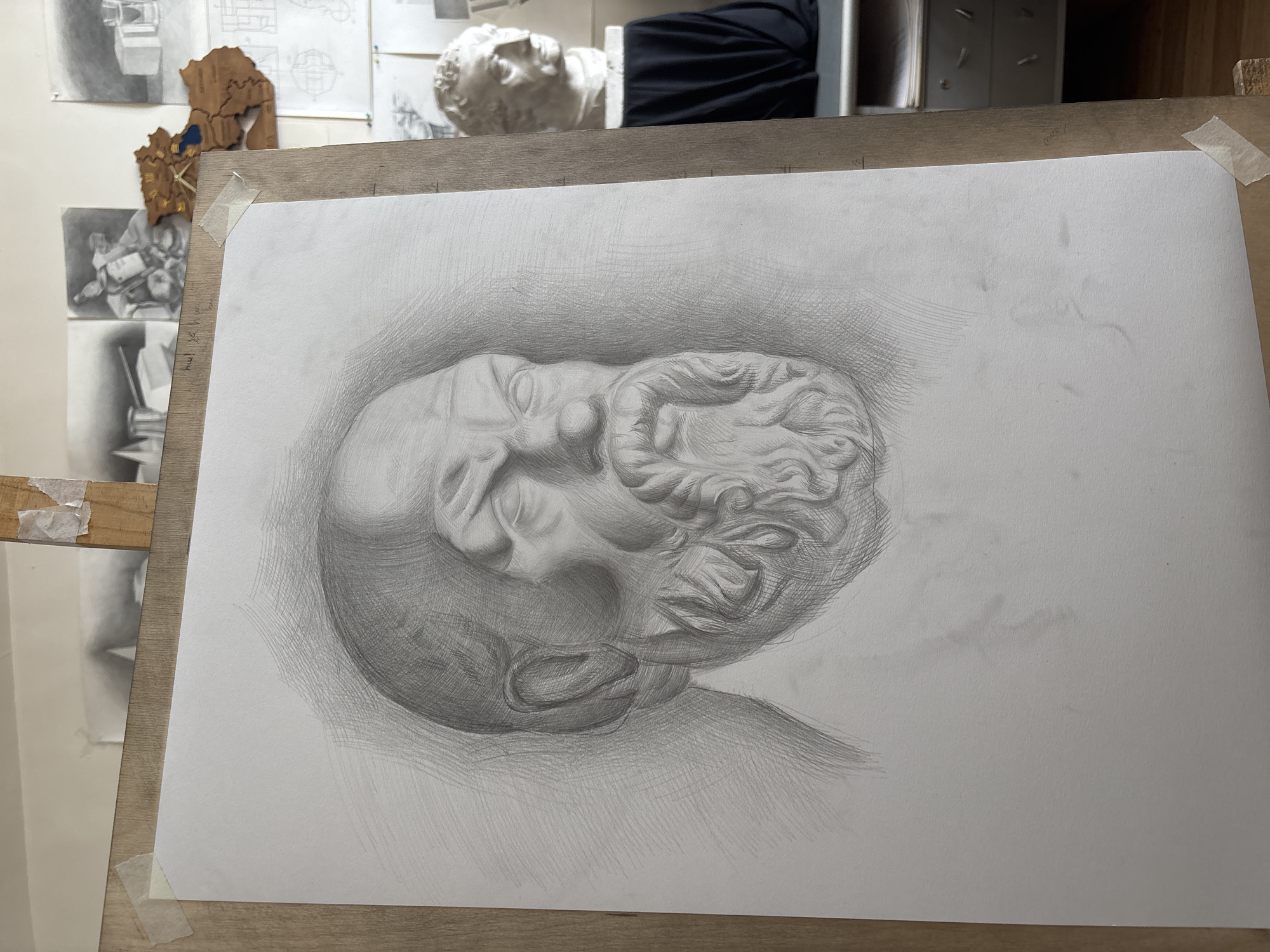 This media file presents a detailed pencil drawing of a classical plaster bust, likely depicting a bearded male figure such as Hercules or Zeus, in an art studio setting in Yerevan, Armenia. The drawing, executed with careful shading, hatching, and cross-hatching techniques, captures the form, light, and shadow of the bust. The figure is shown from a dynamic, slightly low angle, looking upwards and to the right, with a softly shaded background creating a vignette effect around the head and neck.
The drawing is affixed with adhesive tape to a light-colored wooden drawing board or easel, which dominates the foreground. In the background, behind the drawing board, an art studio environment is evident. A white plaster bust, seemingly the model for the drawing, is visible on a surface. Other monochrome sketches and drawings are displayed on a wall, along with a unique wooden decorative piece that resembles a stylized map or crest, featuring blue and gold elements. A dark fabric is also partially visible behind the plaster bust. No discernible text is present in the image. The scene suggests an artistic study or a work-in-progress.