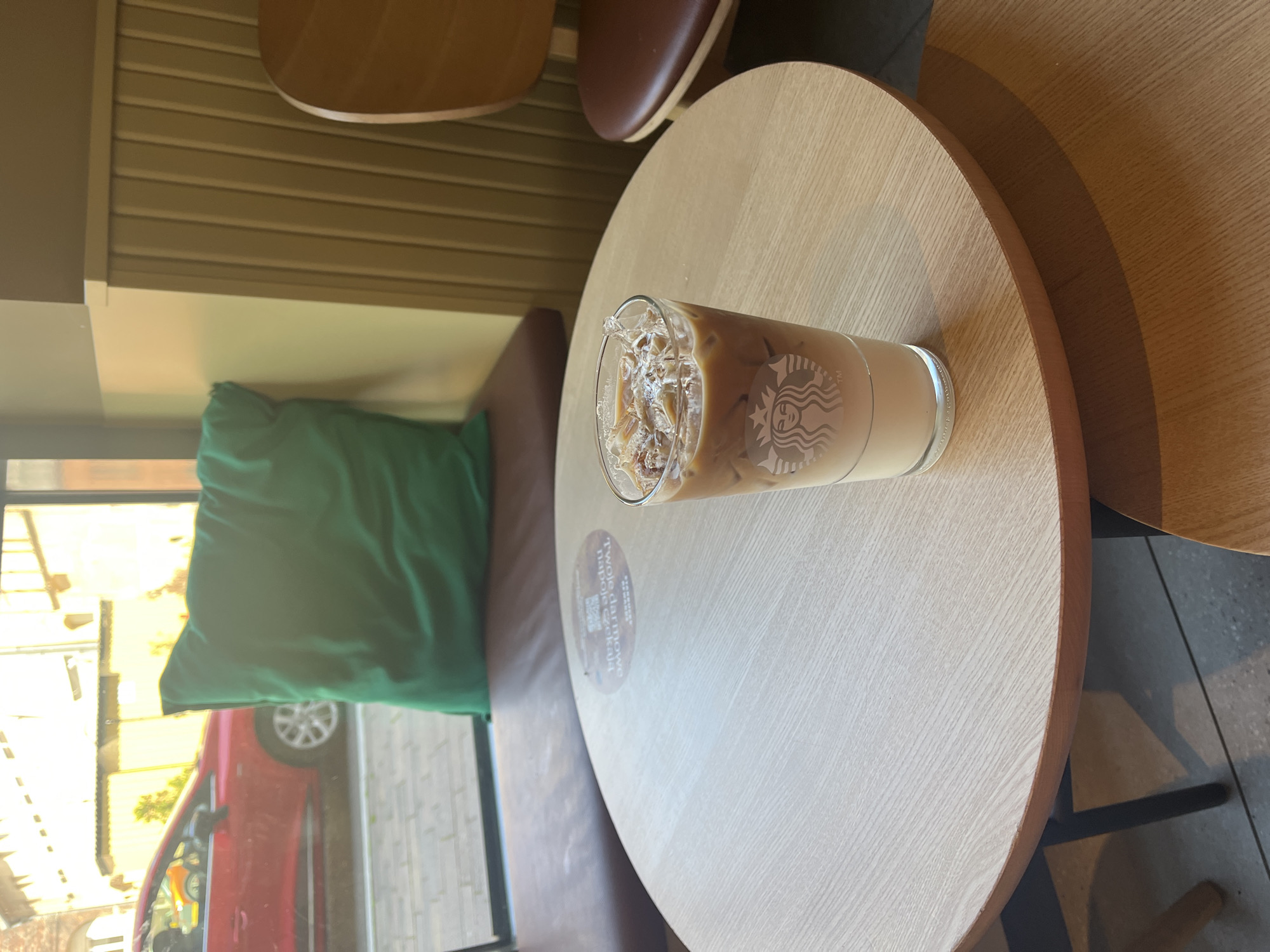 An iced coffee in a Starbucks branded glass sits on a round, light-colored wooden table. The coffee is layered with milk at the bottom and a lighter brown liquid above, topped with ice and some lighter-colored speckles. The Starbucks logo is visible on the glass. A circular decal with text and a QR code is on the table near the glass. In the background, a red car is visible through a window, along with buildings and a bright sky. A large, green cushion rests on a brown upholstered bench by the window. A wooden chair with a brown seat is partially visible to the right, and another smaller wooden table is in the foreground. The environment appears to be an indoor seating area, possibly a cafe or restaurant.