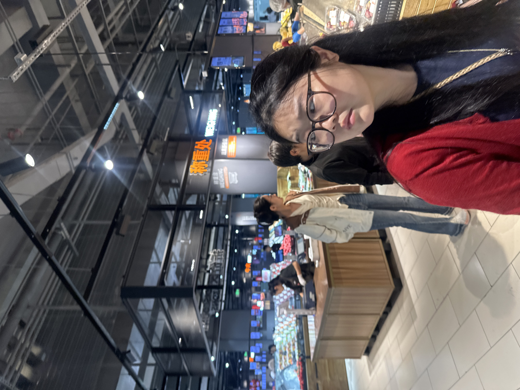 In a brightly lit supermarket in Chongqing High-tech Zone, China, a young woman with long, dark hair and glasses is captured in a close-up shot, looking to her right. She wears a red cardigan over a dark blue shirt and a braided necklace. In the background, other shoppers and supermarket staff are visible. One woman in the mid-ground is wearing a white jacket and blue jeans, and appears to be interacting with a display of produce. Another person in a black shirt stands behind a wooden counter. The environment suggests a modern retail setting with extensive shelving and overhead lighting. The overall scene is consistent with a typical grocery shopping experience.