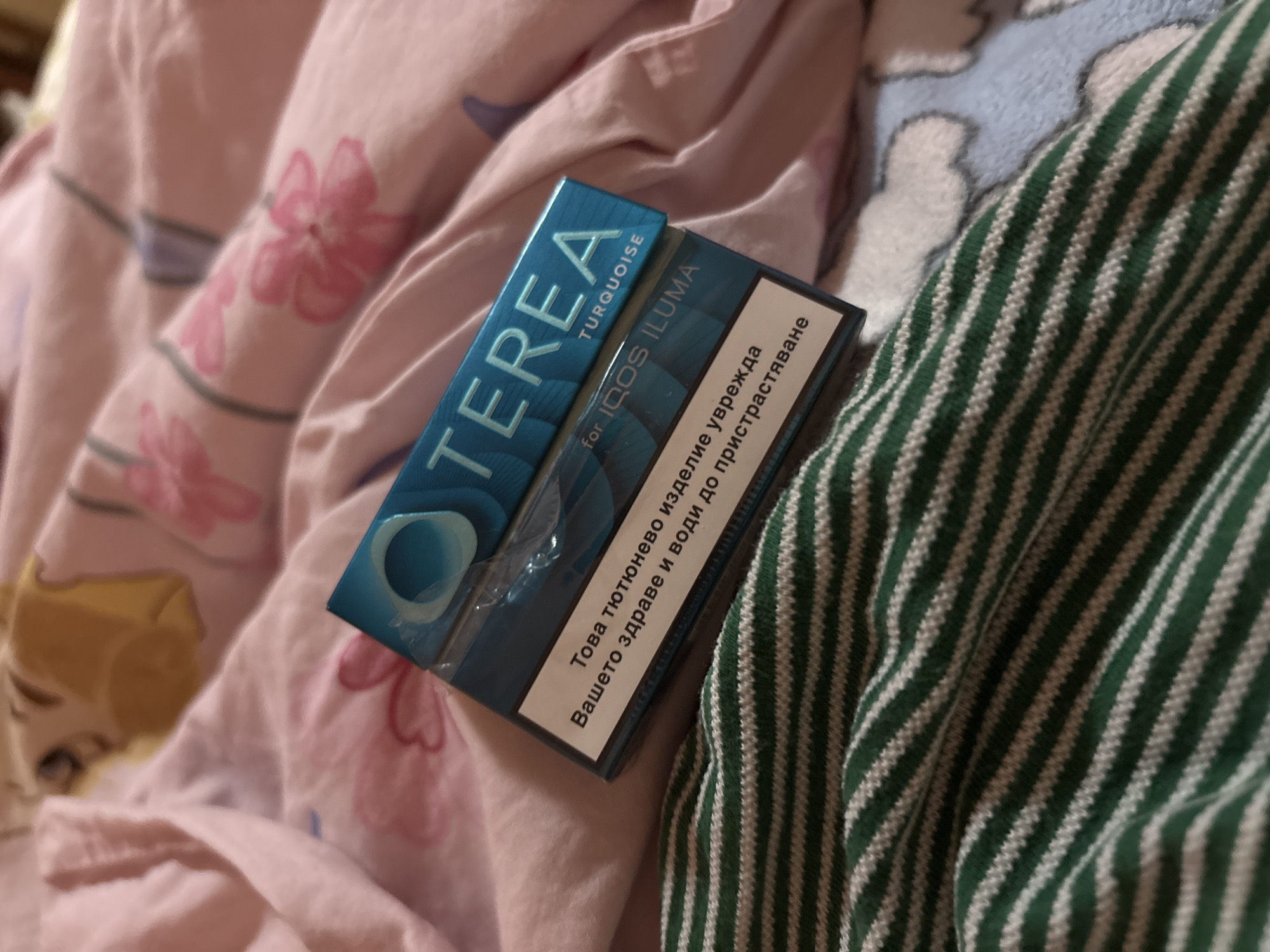 The image shows a pack of "TEREA Turquoise for IQOS ILUMA" cigarettes. The pack rests on a bed with pink floral-patterned sheets and a green and white striped fabric. The location is Garmen, Bulgaria, as indicated by the Bulgarian text on the cigarette pack.