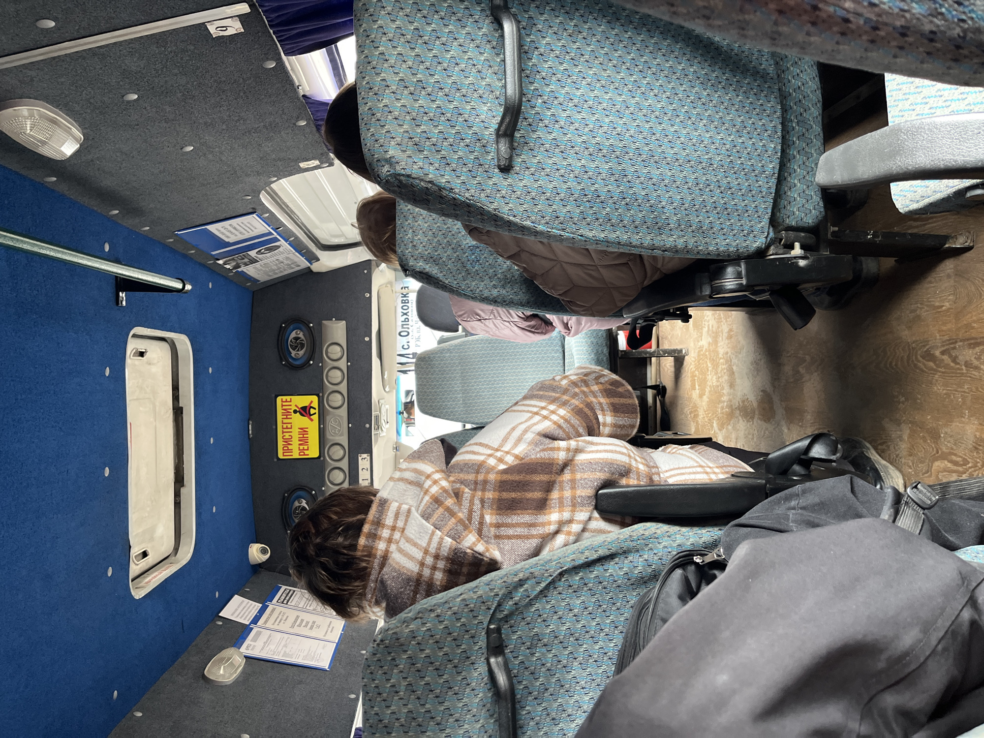 The interior of a passenger vehicle, possibly a van or small bus, is shown. Two passengers are visible. One passenger is draped in a brown and white plaid blanket. The vehicle appears to be in motion, with the passengers seated in blue upholstered seats. A panel with a sign written in Cyrillic that translates to "Fasten Seat Belts" and other indicator buttons is visible. A document, likely a route information, is affixed to the side of the vehicle. Text reading "s. Olkhovka" with accompanying Cyrillic text suggests the location is near or in the village of Olkhovka, which is near Chaikovsky, Russia.