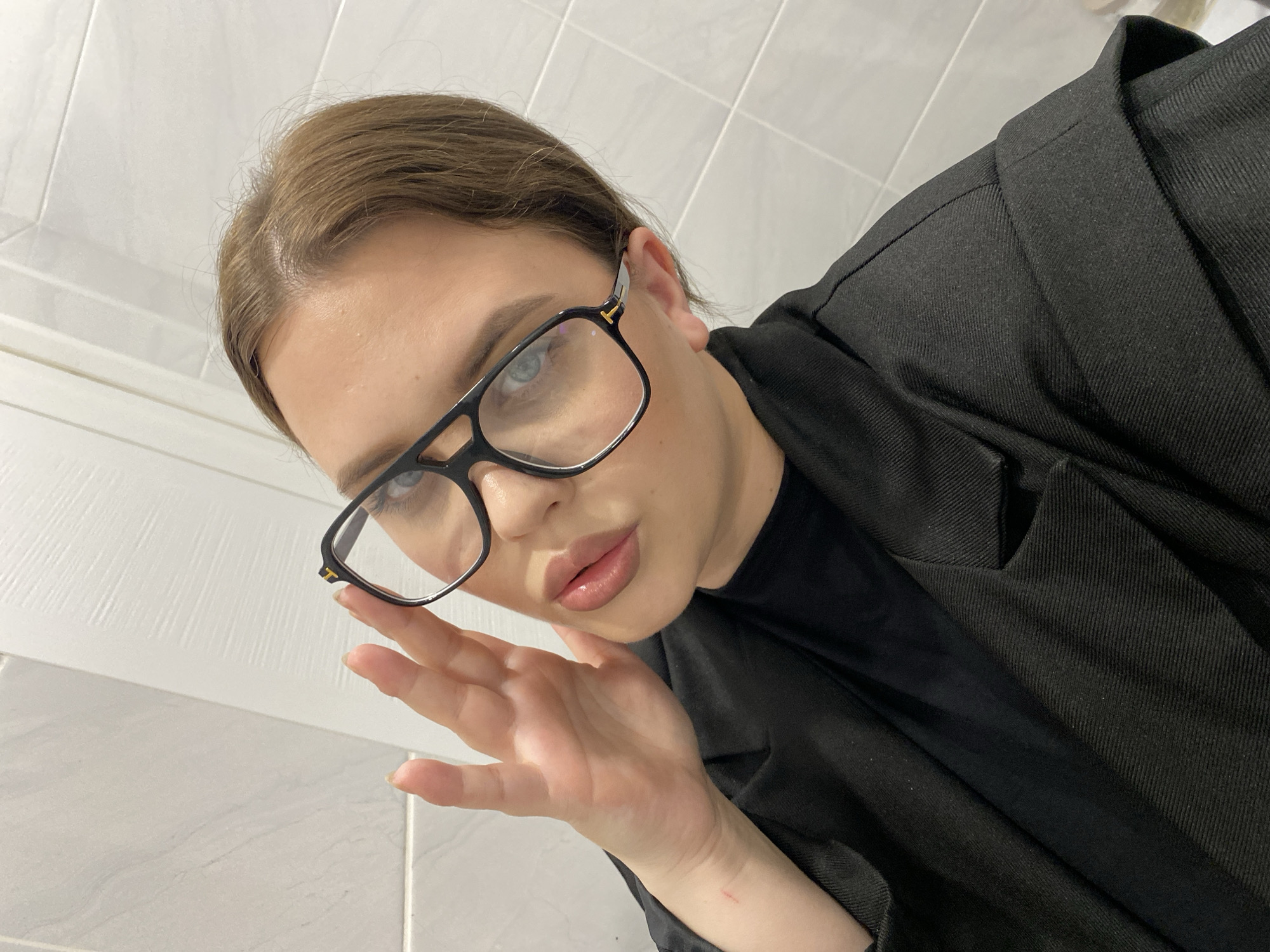 A close-up, high-angle selfie captures a young person, likely female, with light brown hair and light blue-grey eyes, looking directly into the camera. They wear black-framed glasses featuring a small gold 'T' logo on the temples, suggesting a designer brand such as Tom Ford. The individual is dressed in a black blazer or jacket made of a textured, possibly ribbed, fabric, worn over a black top. Their lips are slightly parted in a soft pout, and one hand is raised near their chin, with fingers gently curled. A faint red scratch is visible on the wrist of the raised hand. The background consists of a clean, white tiled wall, implying an indoor setting, possibly a bathroom or kitchen. The lighting suggests an indoor environment. The image conveys a self-assured and stylish demeanor. The location context for this scene is Chernushka, Russia.