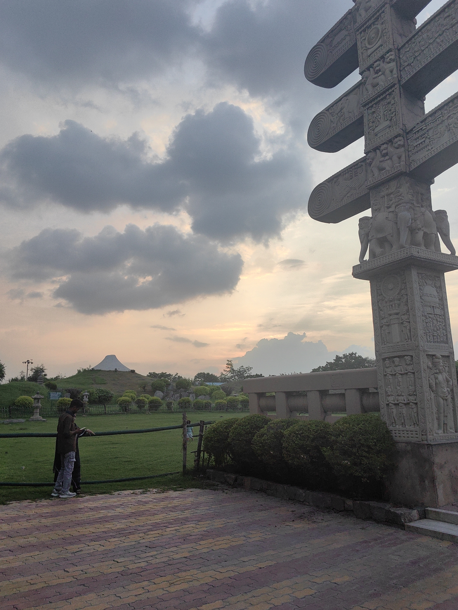 The image captures a scene at the Sanchi Stupa, located in Delhi, India, at dusk. In the foreground, a stone pillar with intricate carvings stands tall, featuring depictions of elephants and floral patterns. To the left, two individuals, one wearing a dark shirt and light-colored pants, the other partially obscured by a dark garment, stand near a green grassy lawn separated by a dark railing. In the mid-ground, a series of decorative bushes and low walls are visible, leading to a white conical structure on a hill in the background. The sky above is filled with dramatic clouds, with the soft glow of the setting sun filtering through. The ground in the immediate foreground is paved with colorful brickwork.
