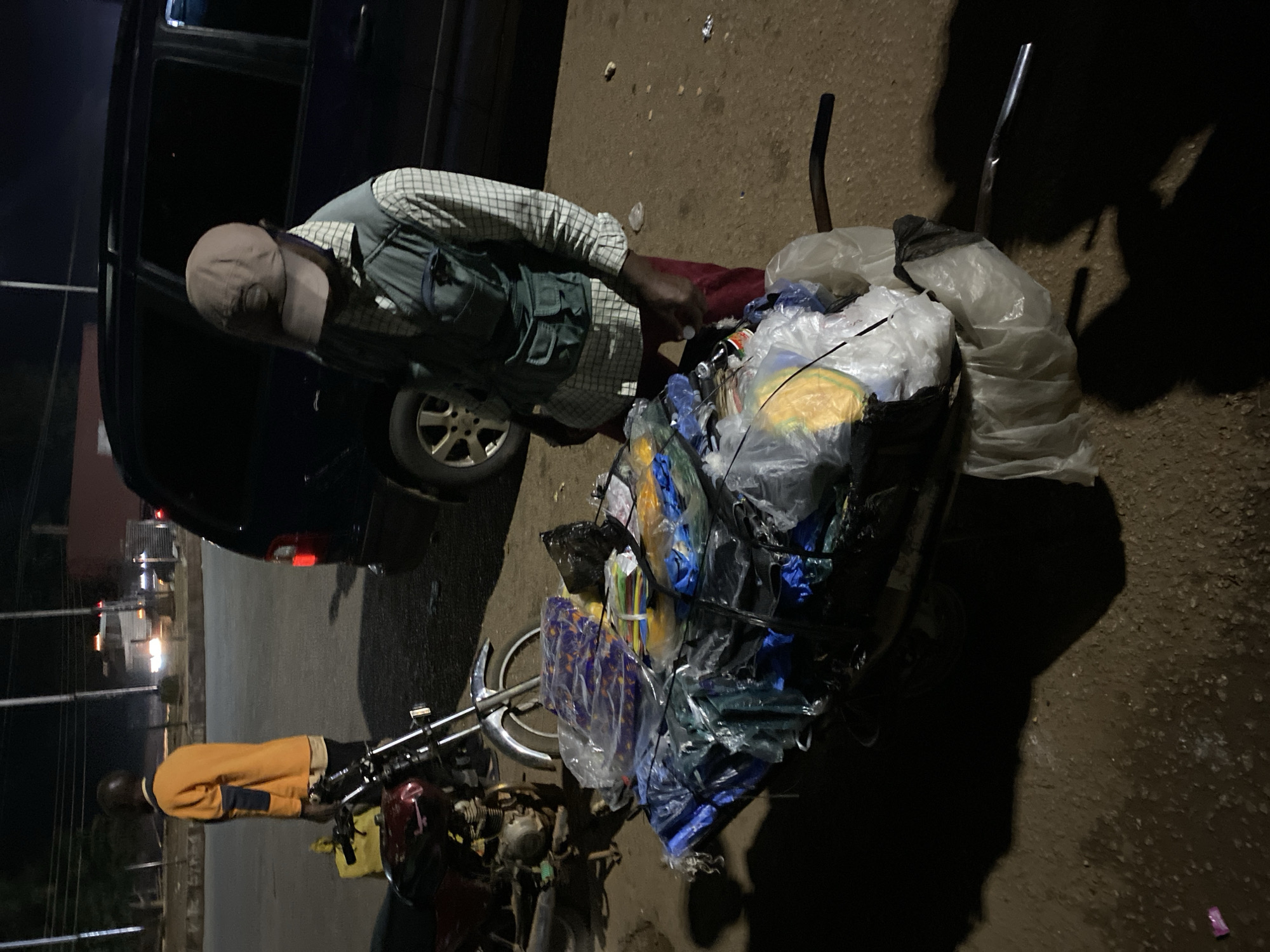 In the city of Jos, Nigeria, at nighttime, a man wearing a light brown baseball cap, a checkered long-sleeved shirt, and a green vest with multiple pockets stands next to a cart piled high with various items, primarily wrapped in plastic. The cart is attached to the back of a motorcycle. The motorcycle's front wheel and part of its frame are visible, as is a maroon-colored fuel tank. In the background, a dark blue SUV is parked. Another person, wearing an orange long-sleeved shirt and dark trousers, is standing further back, facing away from the camera. The ground appears to be unpaved or dusty. The scene is illuminated by artificial lights, casting strong shadows.