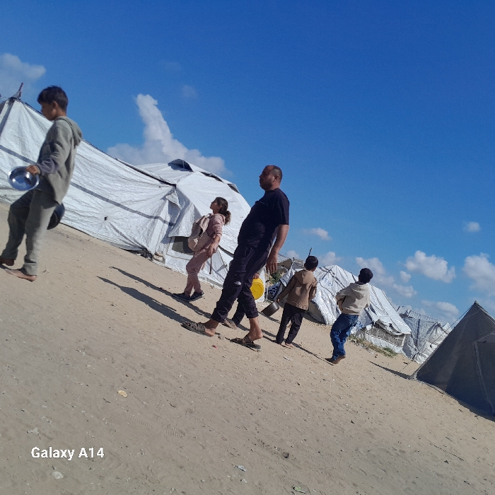 Five individuals walk across sandy terrain under a clear blue sky in Khan Yunis, Palestinian Territory. In the background, numerous white tarpaulin tents, some appearing disheveled, form a makeshift camp. A darker, conical tent is visible on the far right. The ground is dry and uneven, sloping upwards from left to right. Strong shadows are cast to the left of the individuals, indicating direct sunlight.
From left to right:
1. A young male in a grey hooded tracksuit walks with a silver bowl in his left hand and a dark cylindrical object in his right, his feet bare.
2. A young female in a light pink tracksuit with a light-colored backpack on her shoulders walks with her head tilted upwards. She wears dark shoes.
3. An adult male with a receding hairline, wearing a dark short-sleeved t-shirt, dark cargo pants, and light-colored slide sandals, carries a yellow bucket in his left hand.
4. A young male in a light brown hooded jacket and dark pants carries a white bucket in his left hand.
5. A young male in a light-colored hooded top and blue jeans walks towards the right.
The sky is bright blue with scattered white clouds. A "Galaxy A14" watermark is present in the lower-left corner.