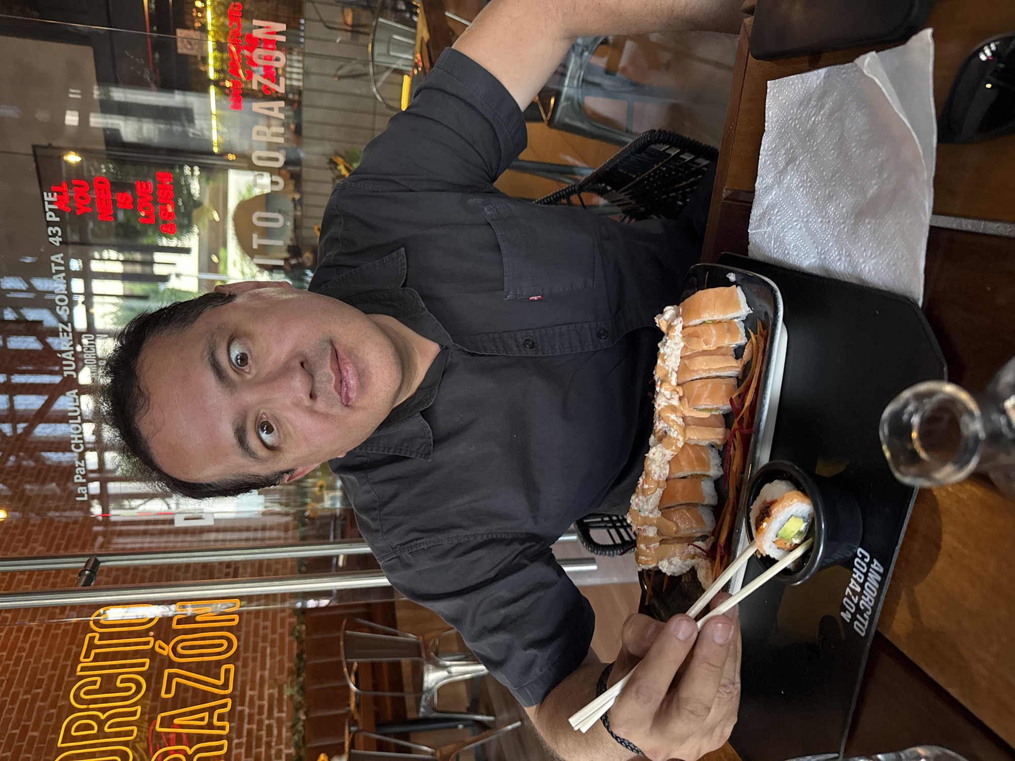 A man in a black button-down shirt is holding chopsticks and eating sushi at "Amorcito Corazón" restaurant. The restaurant is located at Juarez Sonata 43 Pte, La Paz Cholula in San Bernardino Tlaxcalancingo, Mexico. A line of salmon sushi and a separate piece of sushi in a small bowl are visible. A white napkin and a smartphone are on the table. The restaurant's logo, "Amorcito Corazón", is displayed in bright yellow neon lights in the background. The window displays the words "All You Need is Love and Sushi."
